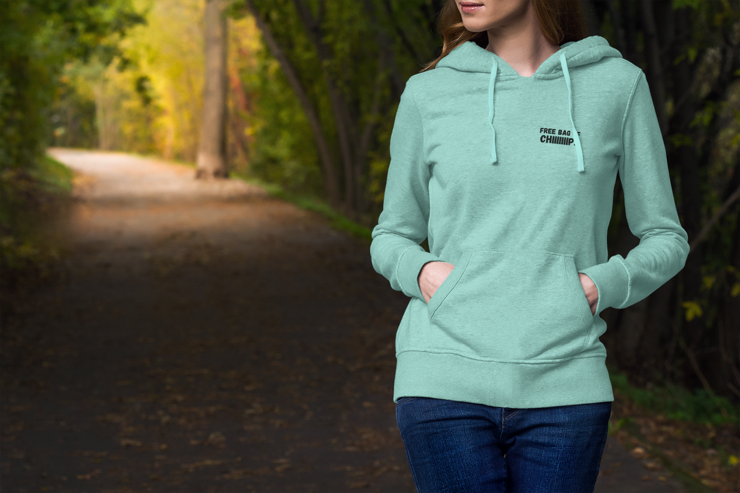 Model vibing in seafoam green "FREE BAG OF CHIIIIIPS" embroidered hoodie, hands in kangaroo pocket on fall forest path