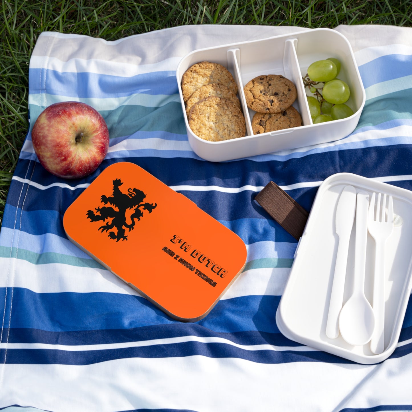 Blue blanket picnic—open white Bammenbak bento loaded with cookies/grapes, orange lion lid, utensils, apple, chocolate bar