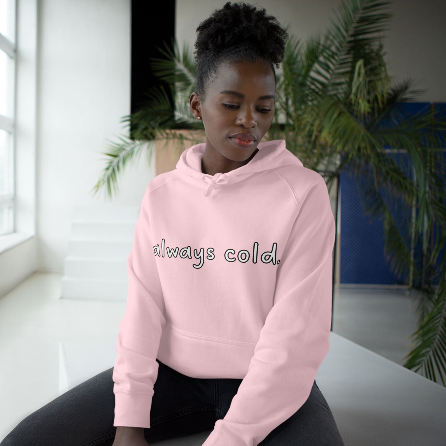 Black model lounging in light pink "always cold." hoodie, front view, white script print on chest