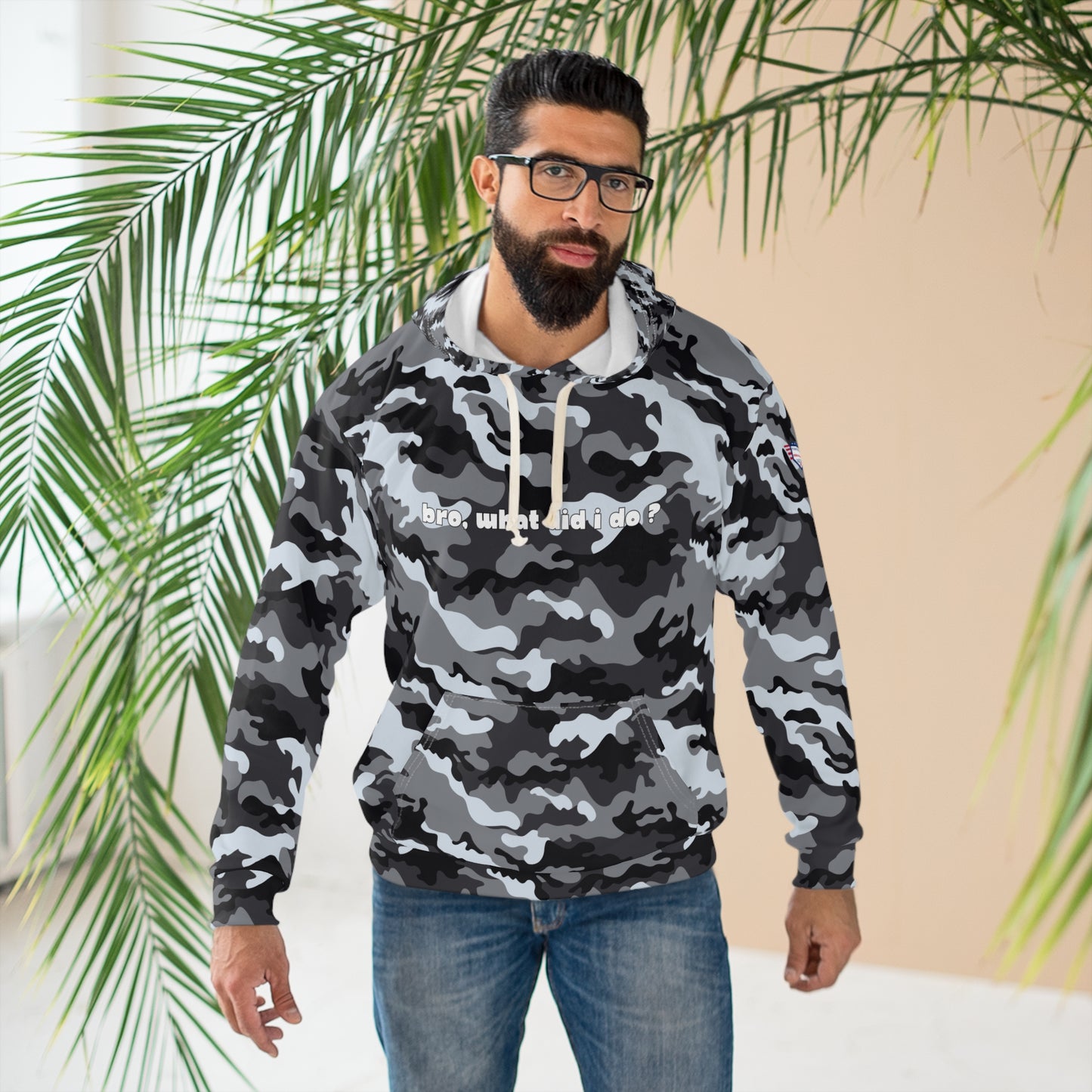 Model with beard and glasses wears grayscale camo hoodie front printed "bro: what did i do?" in white, indoor plant backdrop
