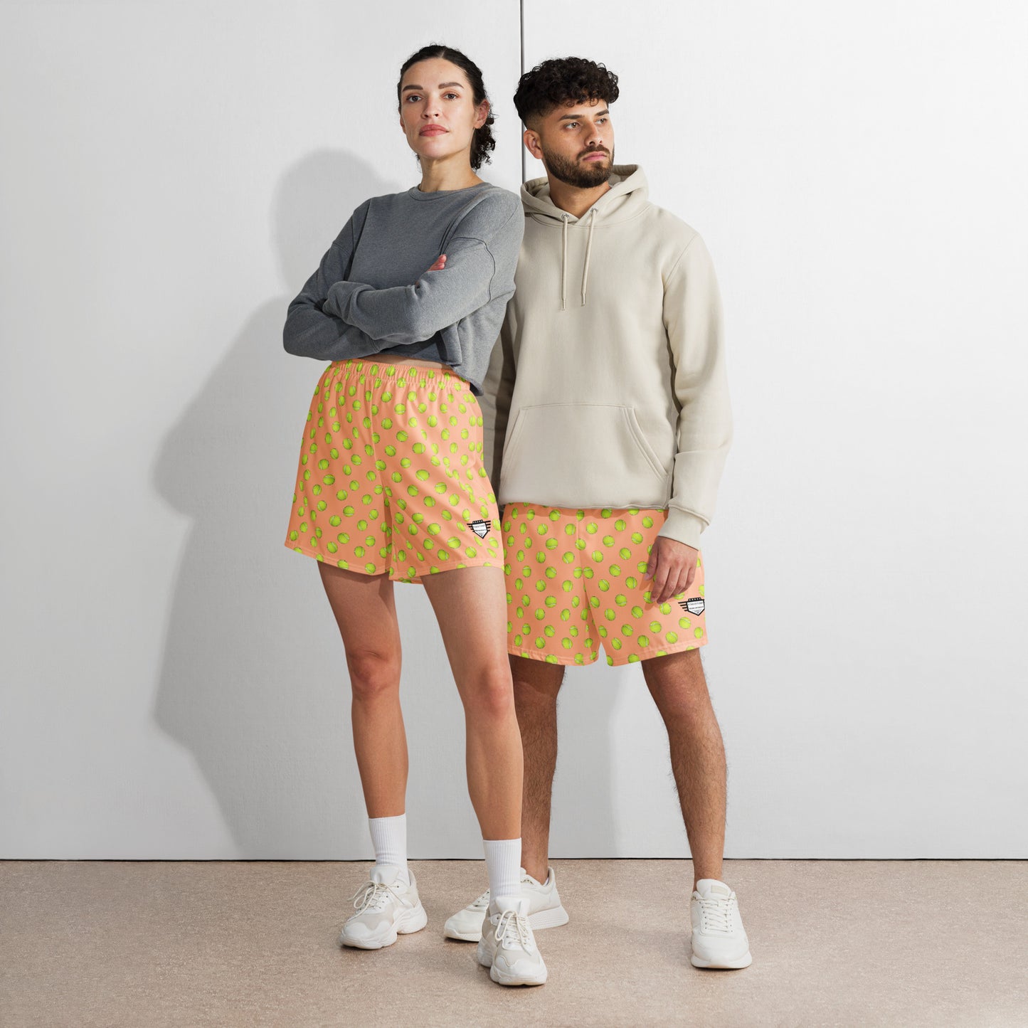 Diverse duo in bright yellow tennis-print athletic long shorts, her in gray hoodie, him in beige — court-ready chill vibes