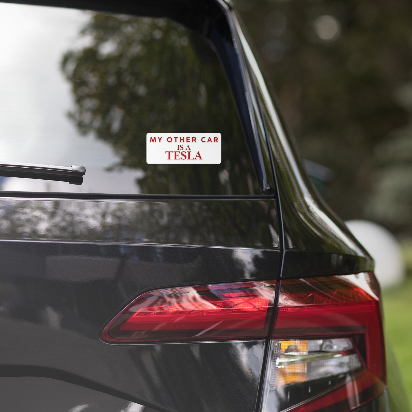 Rear view of black car with white "My other car is a Tesla" sticker on back window, wiper at rest