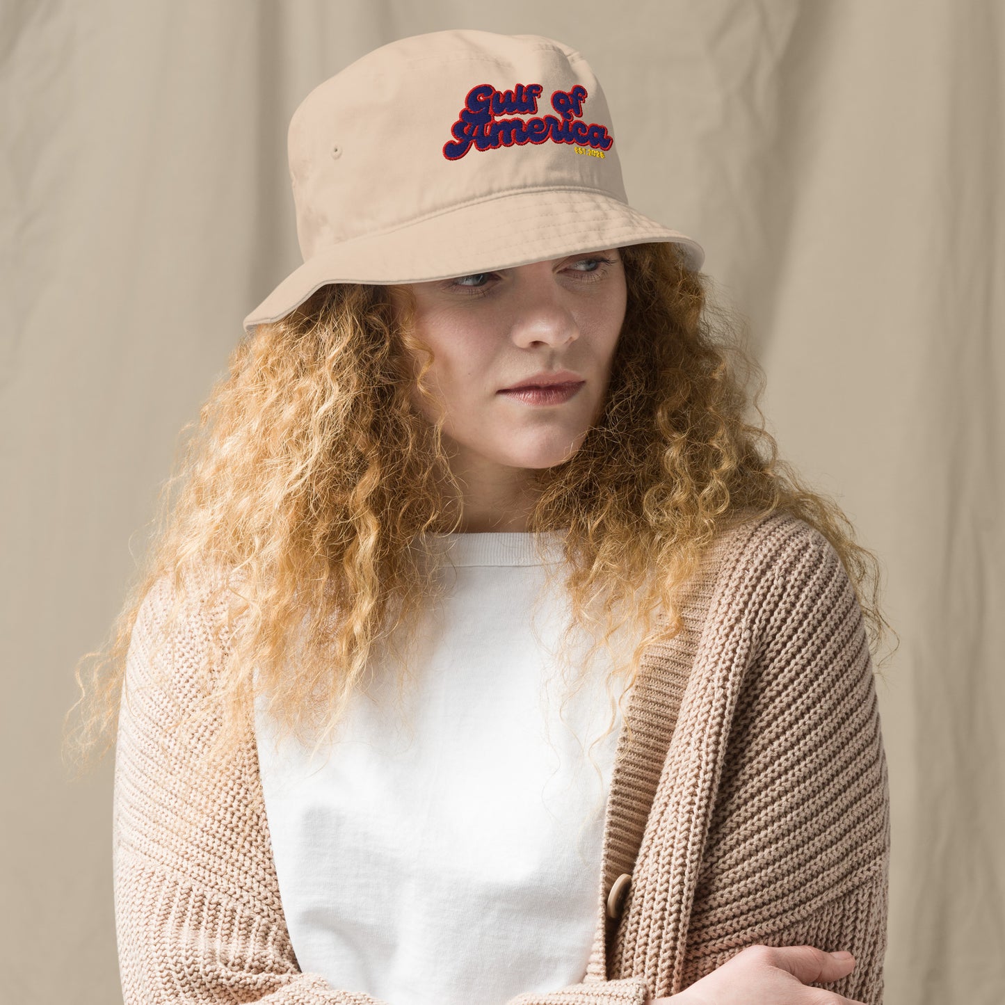 Curly-haired model in three-quarter view wearing beige bucket hat, "Gulf of America" multicolored print front and center