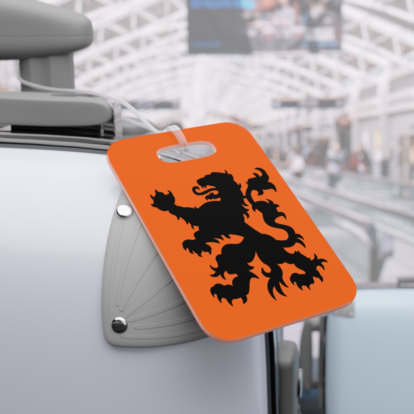 Orange rectangular Dutch Lion tag with black lion attached to gray suitcase handle in airport scene