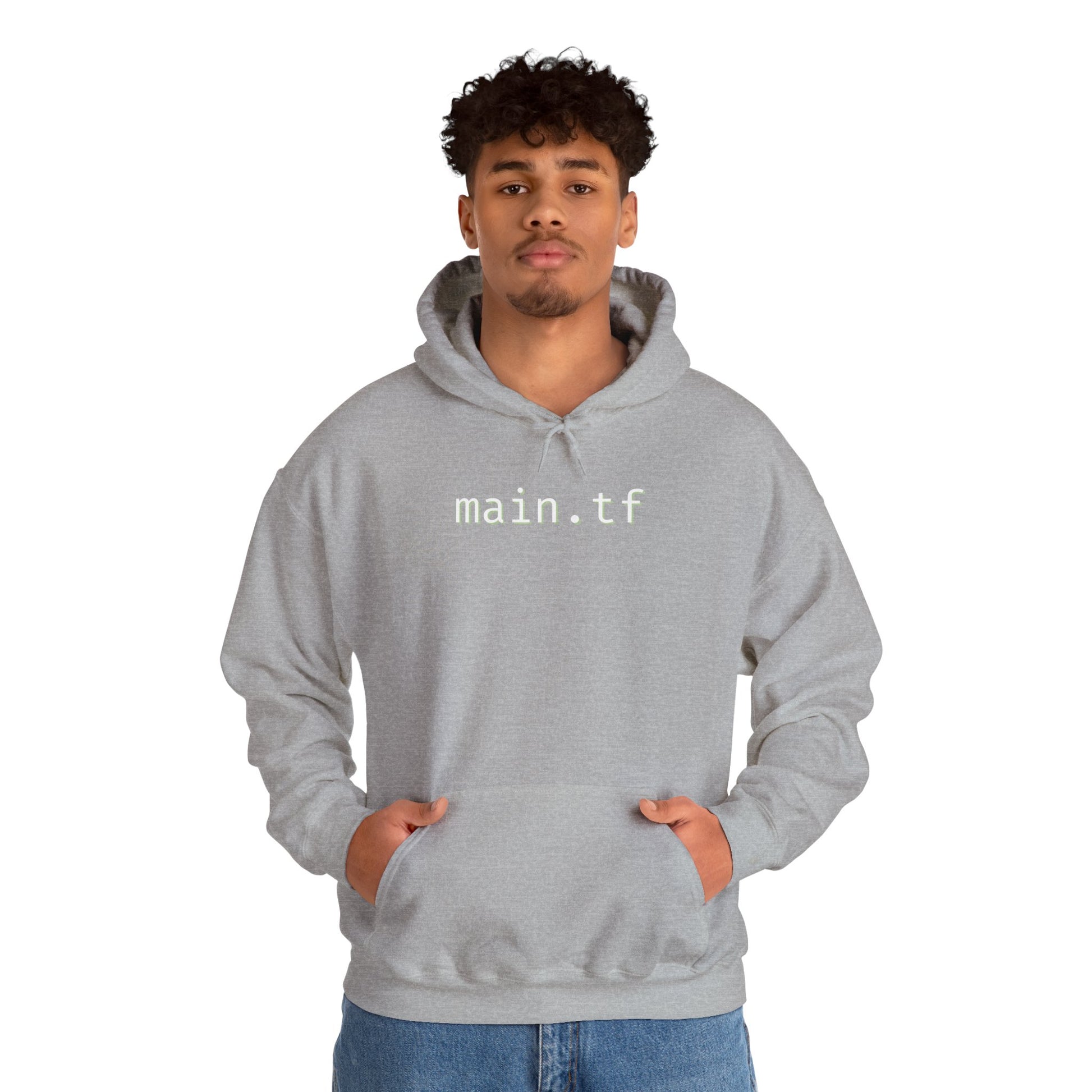 Curly-haired guy in light grey main.tf hoodie, front hands-pocket pose over jeans