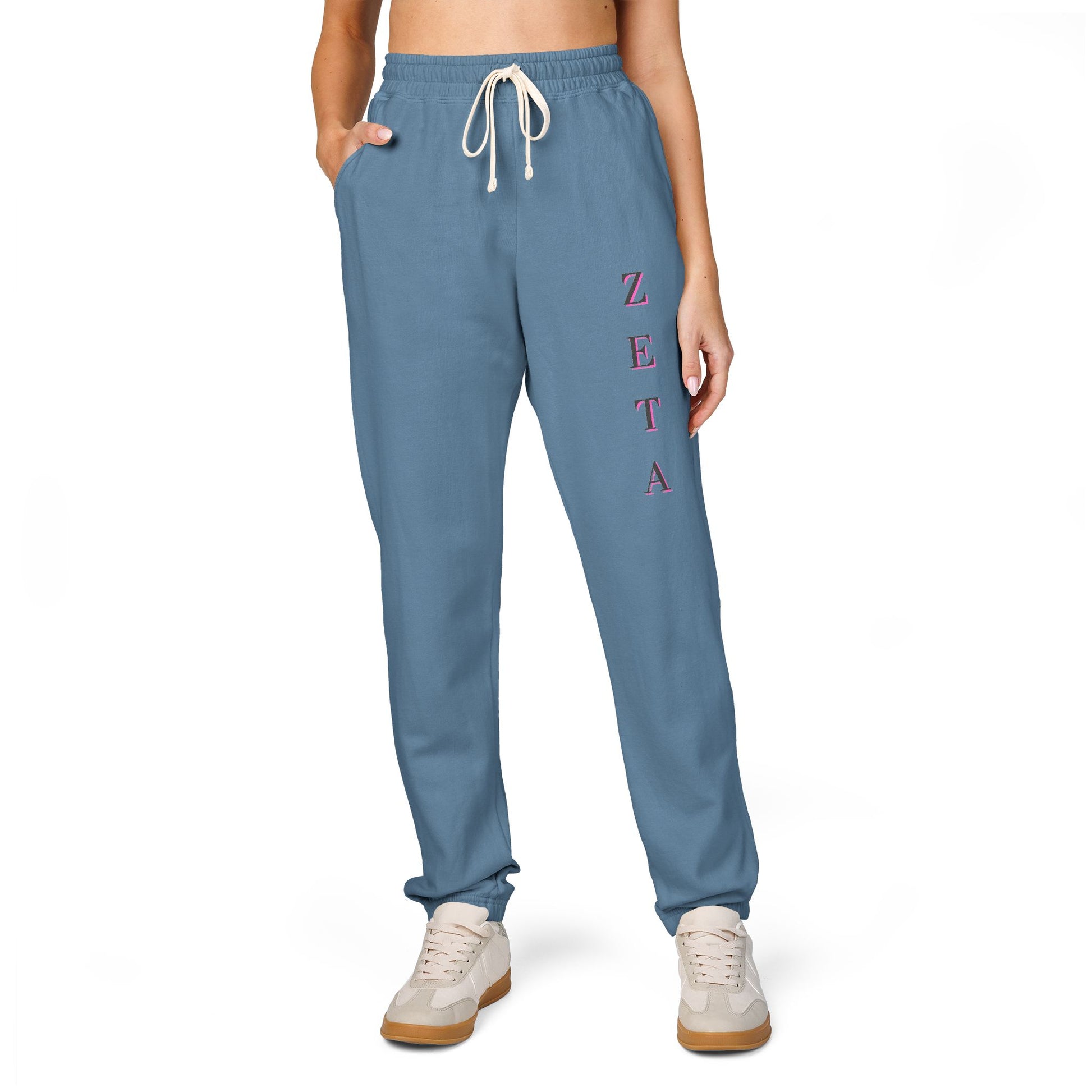 Model strutting front in medium blue Zeta joggers, pink "ZETA" vertical script & white sneakers