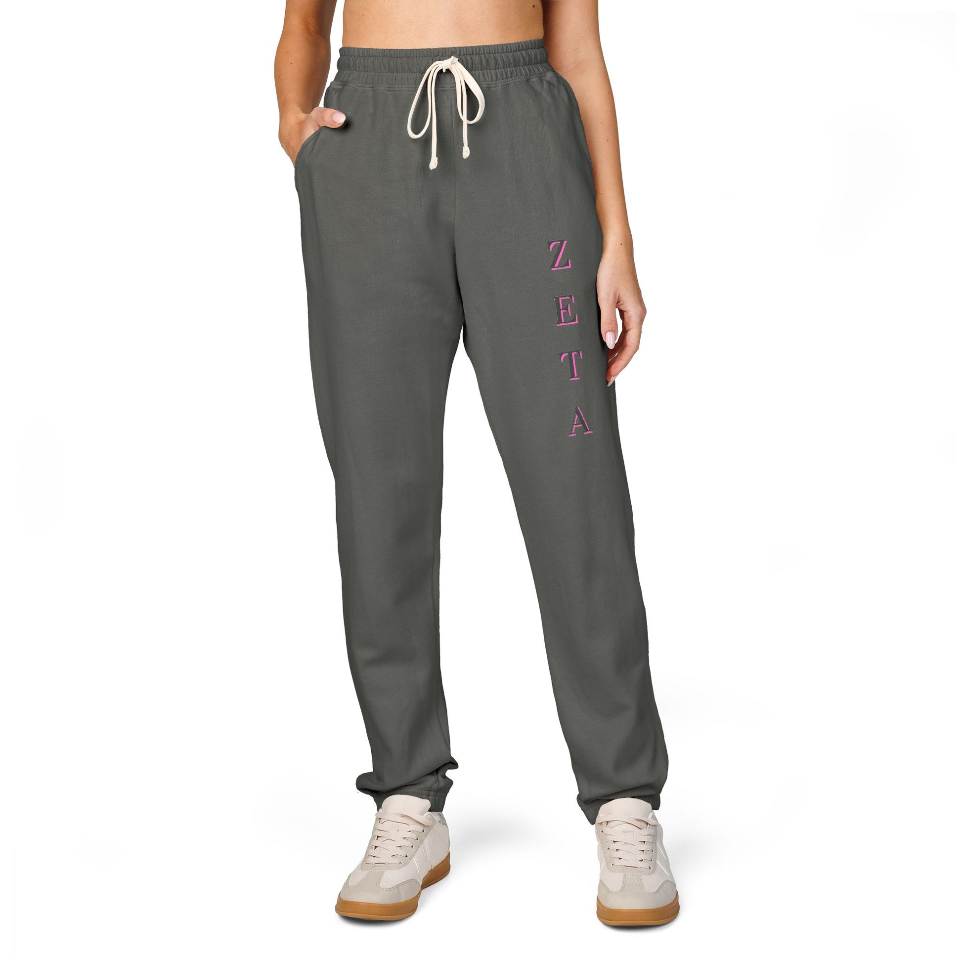 Model front view in charcoal gray Zeta sweatpants, pink vertical "ZETA" script on leg, crop top, white sneakers