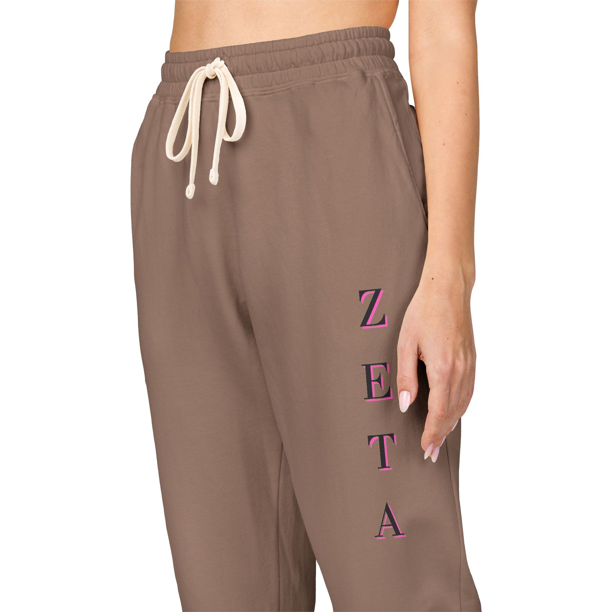 Side close-up of taupe fleece joggers showing pink vertical "ZETA" script, drawstring waist, model's hand