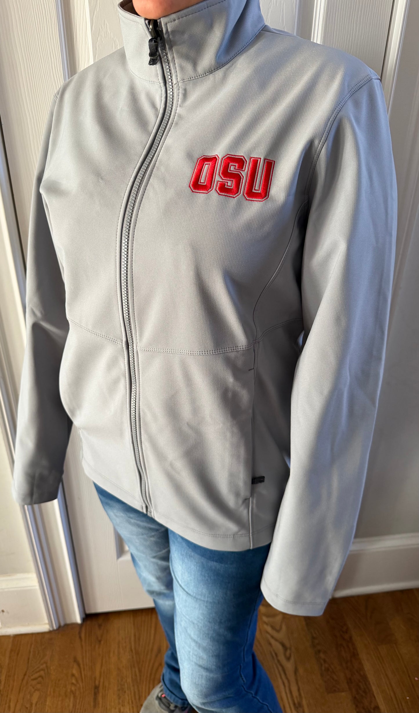 Straight-on front view woman in light grey zipped OSU softshell jacket indoors, red chest logo reps pride