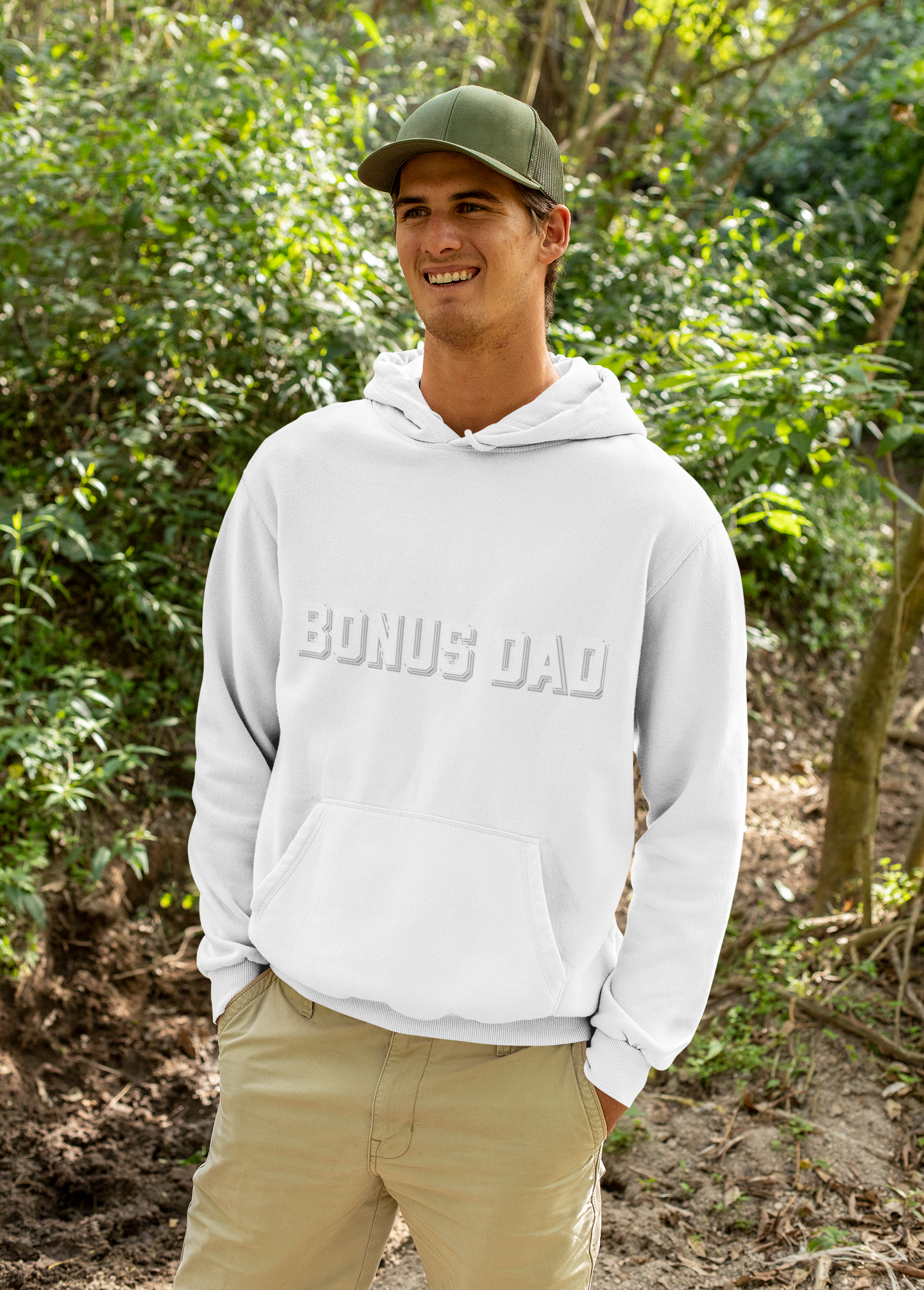 Smiling model rocks front of white "BONUS DAD" embossed hoodie outdoors, green hat and khakis