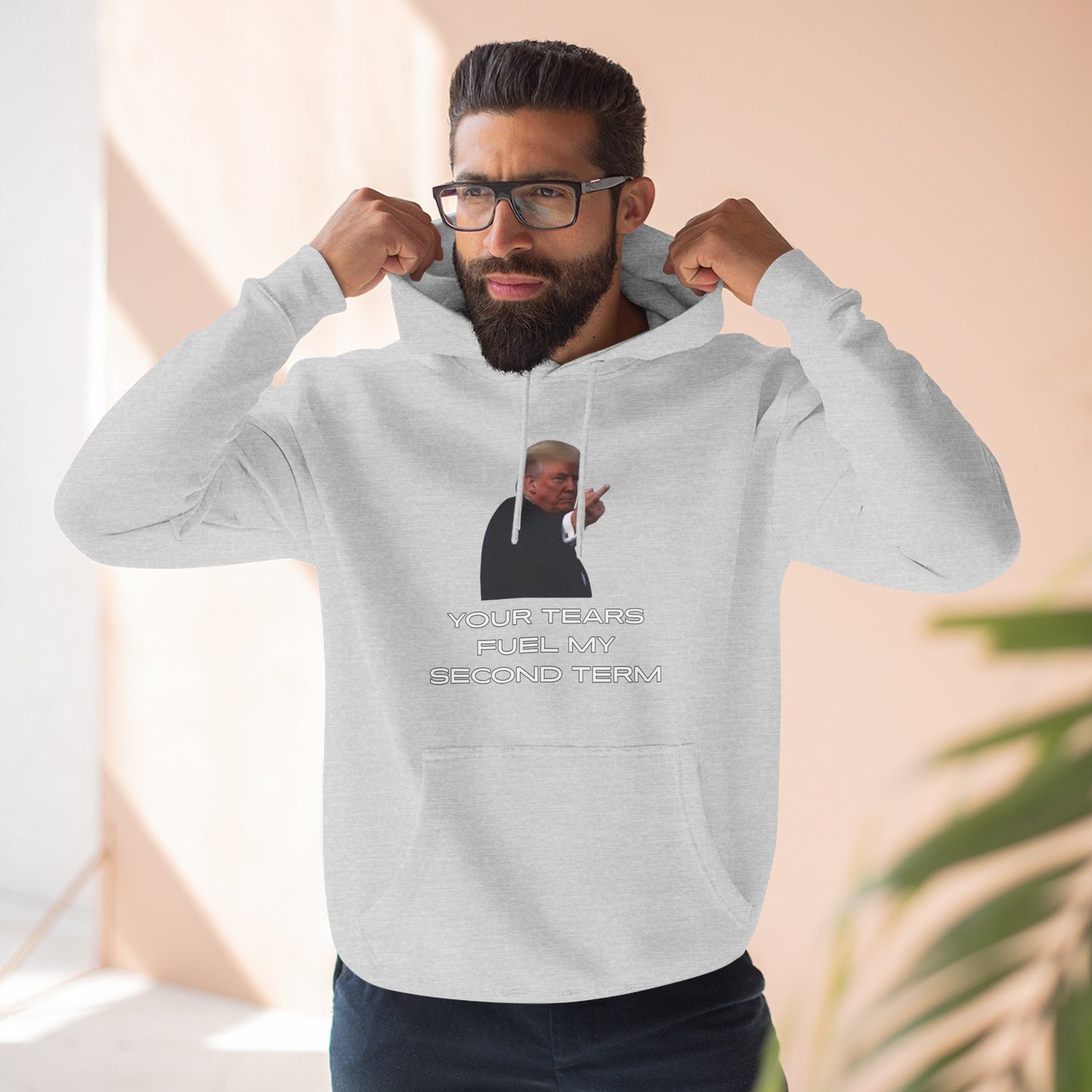 Bearded model flexing arms in heather gray hoodie, front Trump flip-off print with "YOUR TEARS FUEL MY SECOND TERM"