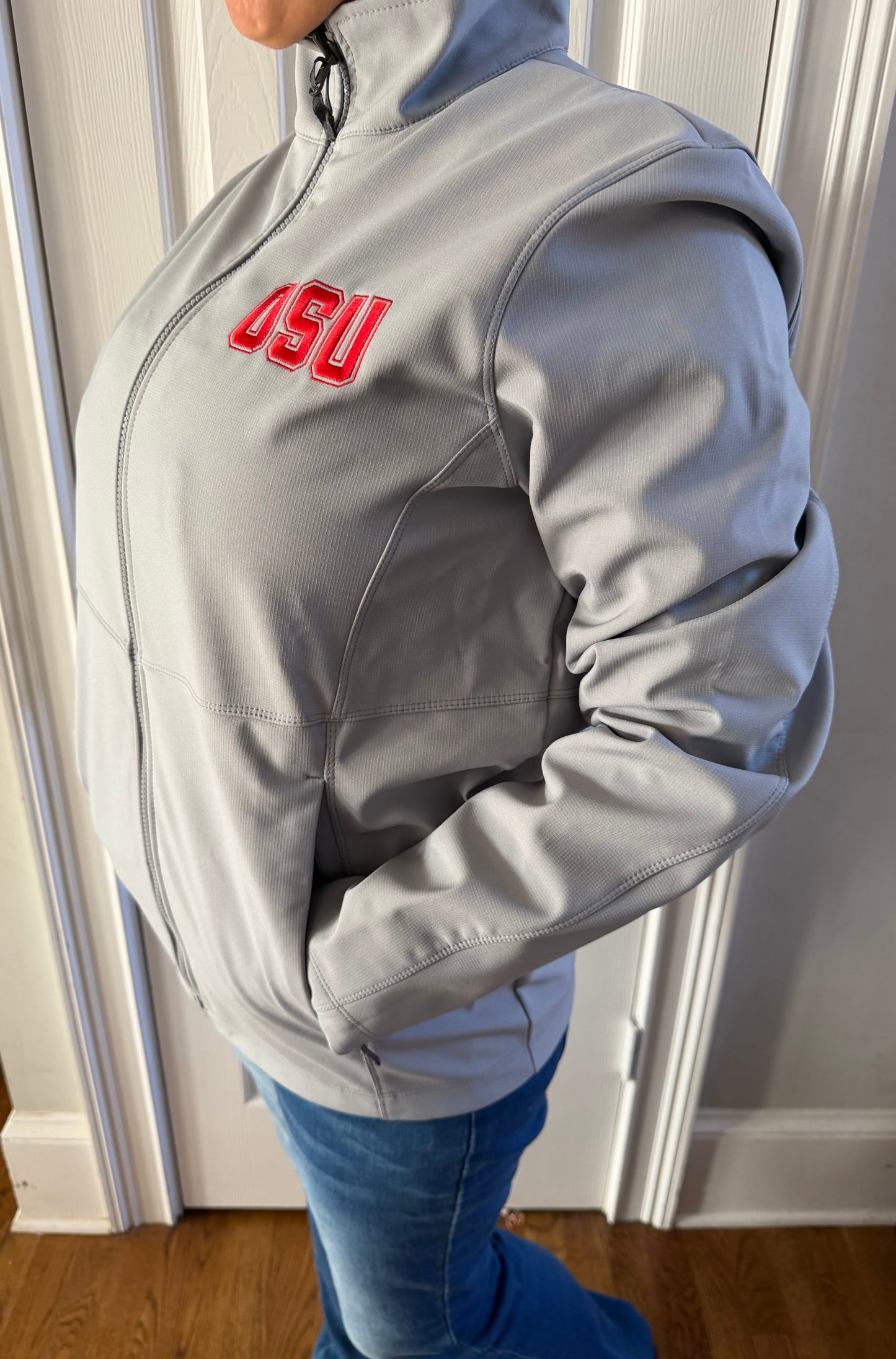 Close front view of woman in light grey full-zip OSU jacket, hands relaxed, red embroidery front and center indoors