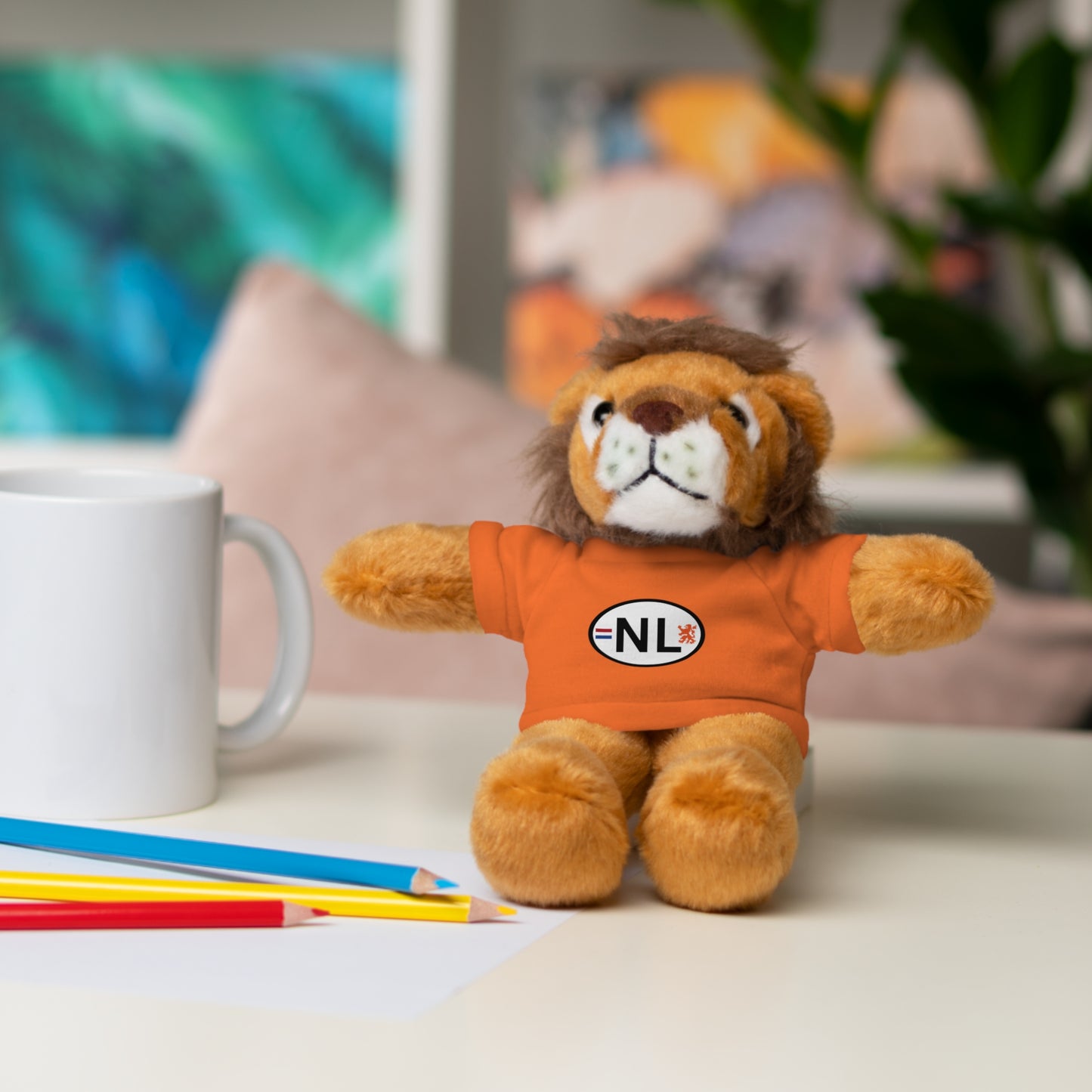 Orange NL lion plush chilling on desk amid pencils, mug and pillows, arms out for World Cup vibes