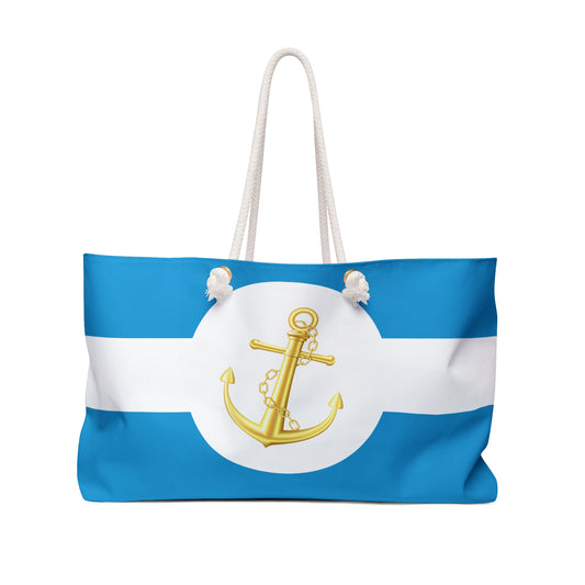 Front view of blue-and-white striped nautical beach tote with shiny gold anchor in white circle, cream rope handles slung high
