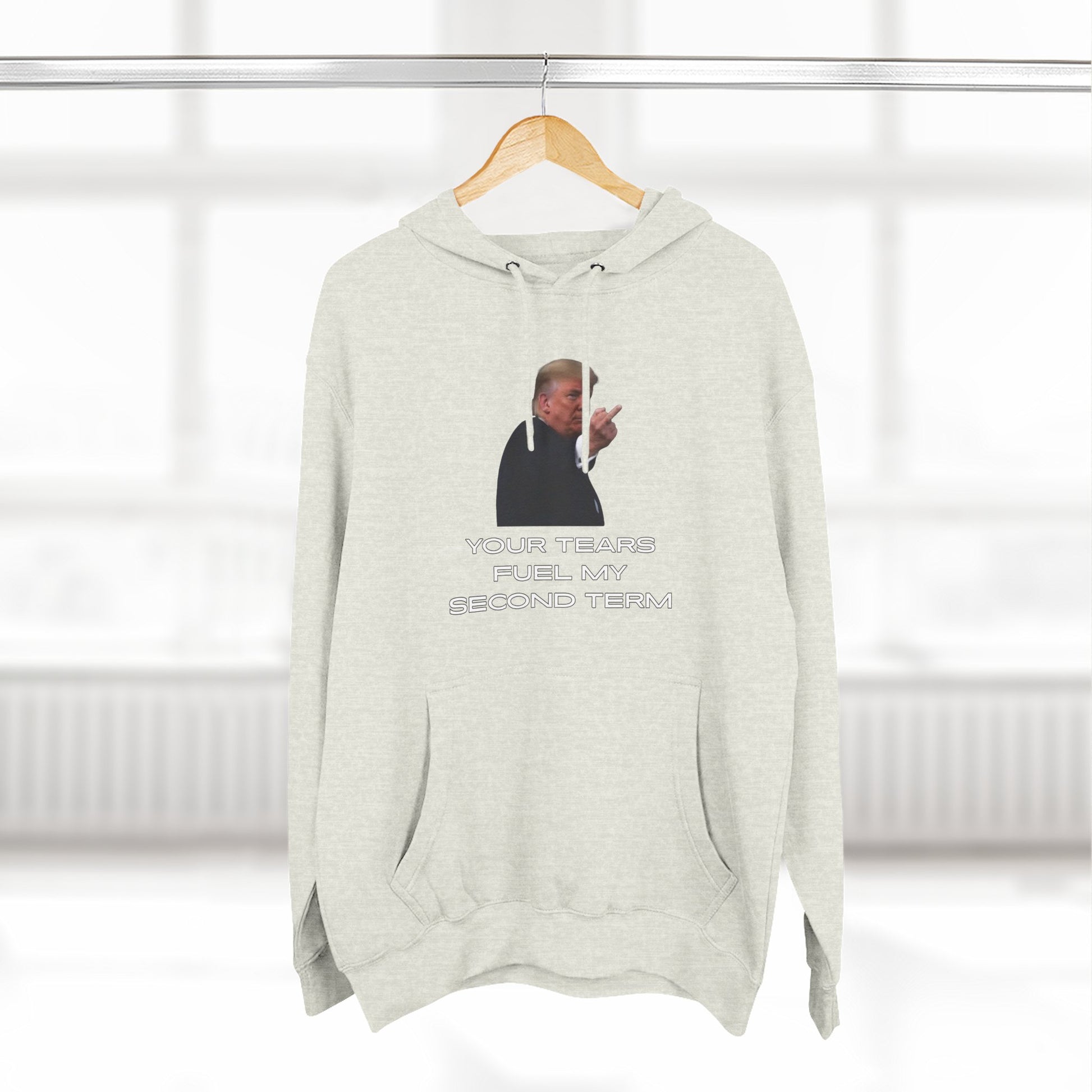 Oatmeal hoodie hanging on rack, front print Trump gesture "YOUR TEARS FUEL MY SECOND TERM"