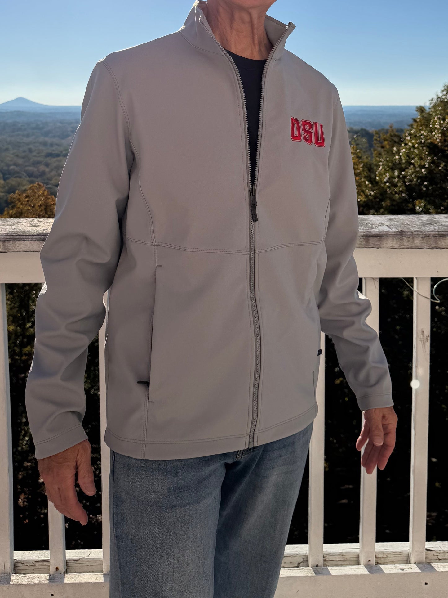 Older man models light grey OSU softshell jacket front-left on sunny balcony, red embroidery pops against mountain backdrop