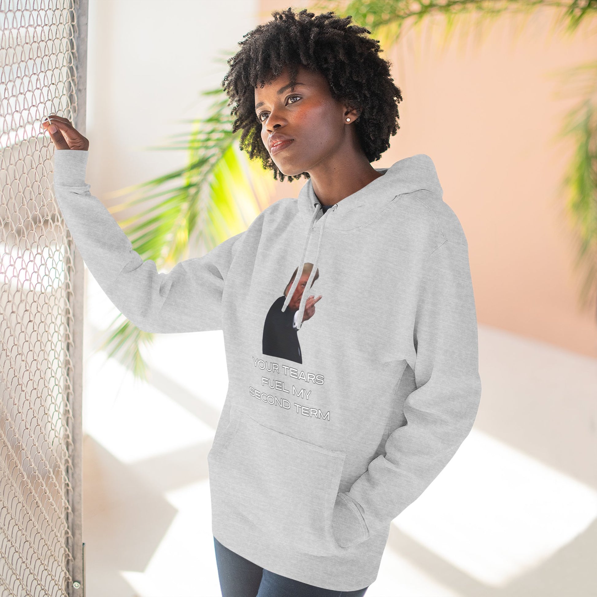 Curly-haired model leaning in heather gray Trump hoodie, front view of middle finger graphic and bold text
