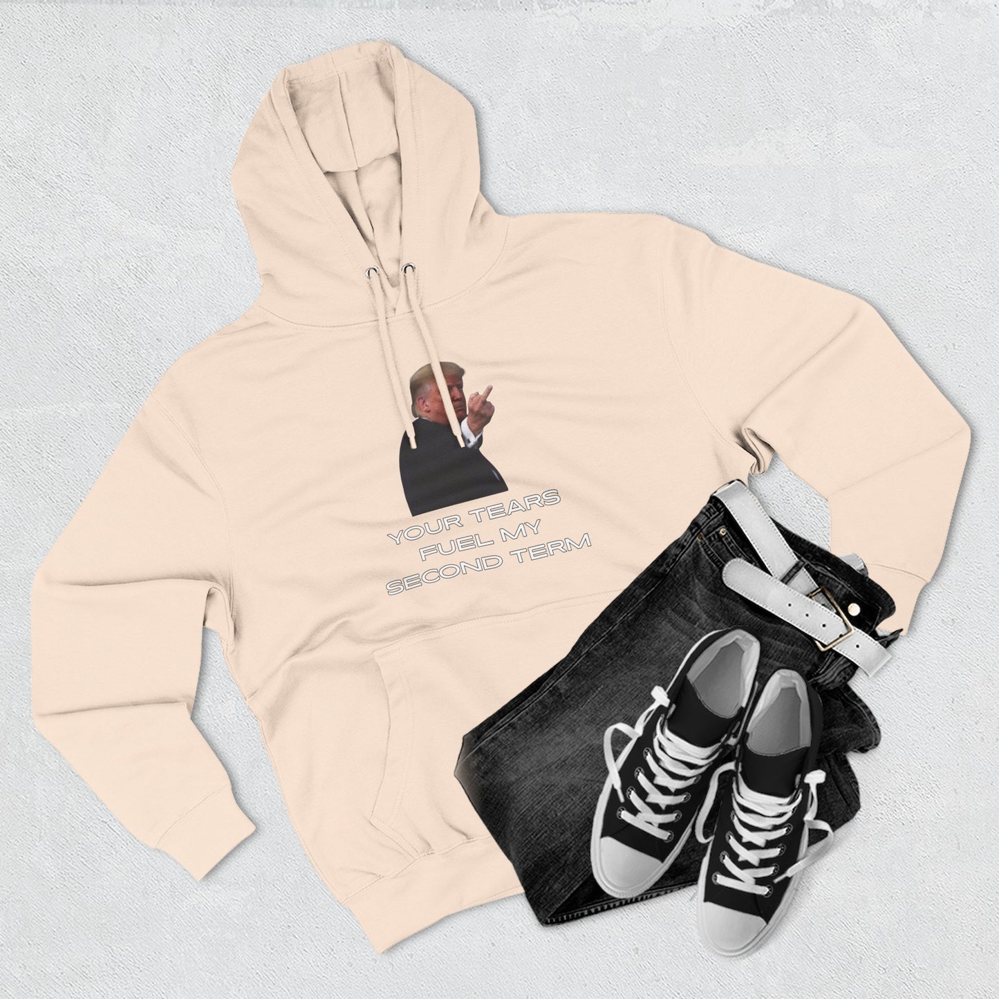 Flat lay oatmeal hoodie front, Trump flip-off print "YOUR TEARS FUEL MY SECOND TERM" beside jeans and sneakers