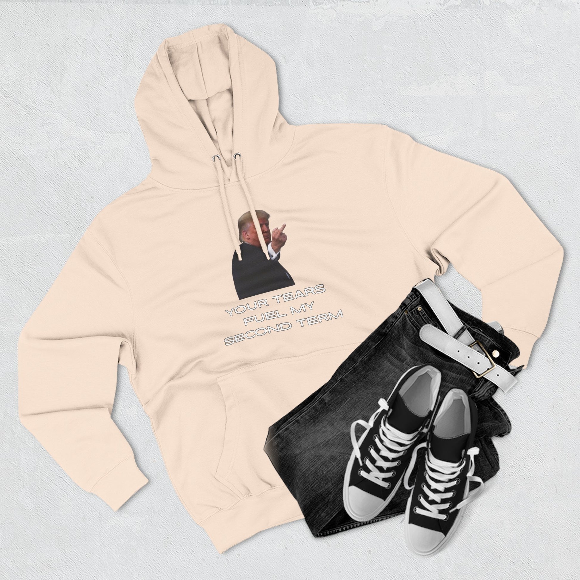 Flat lay oatmeal hoodie front, Trump flip-off print "YOUR TEARS FUEL MY SECOND TERM" beside jeans and sneakers