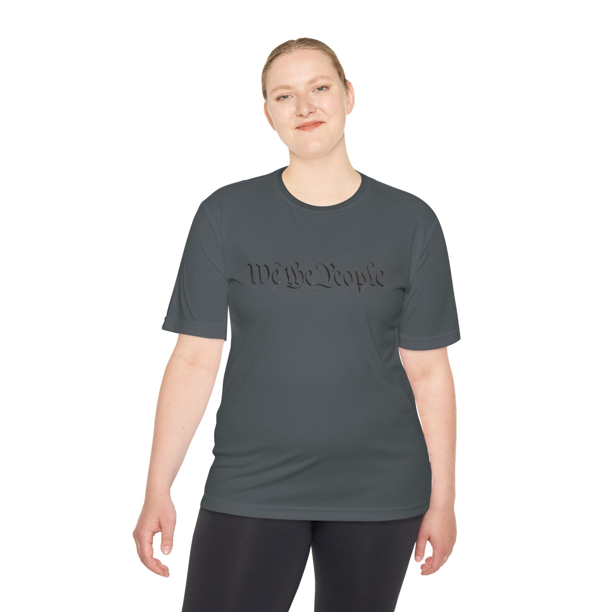 Female model smirking in dark grey moisture-wicking tee, front "We the People" script in black cursive