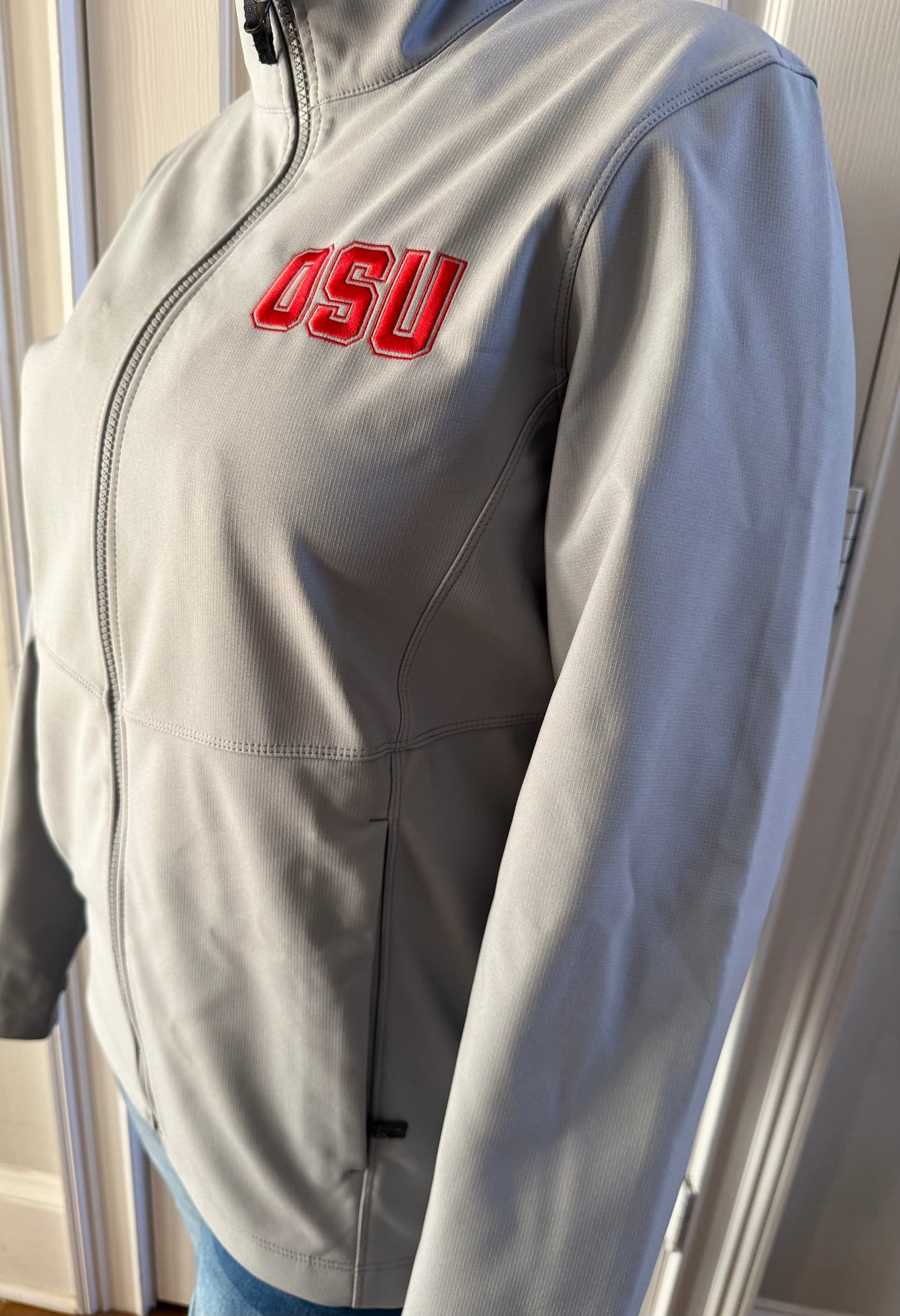 Woman models light grey OSU jacket three-quarter front-right indoors, red embroidery bold on softshell
