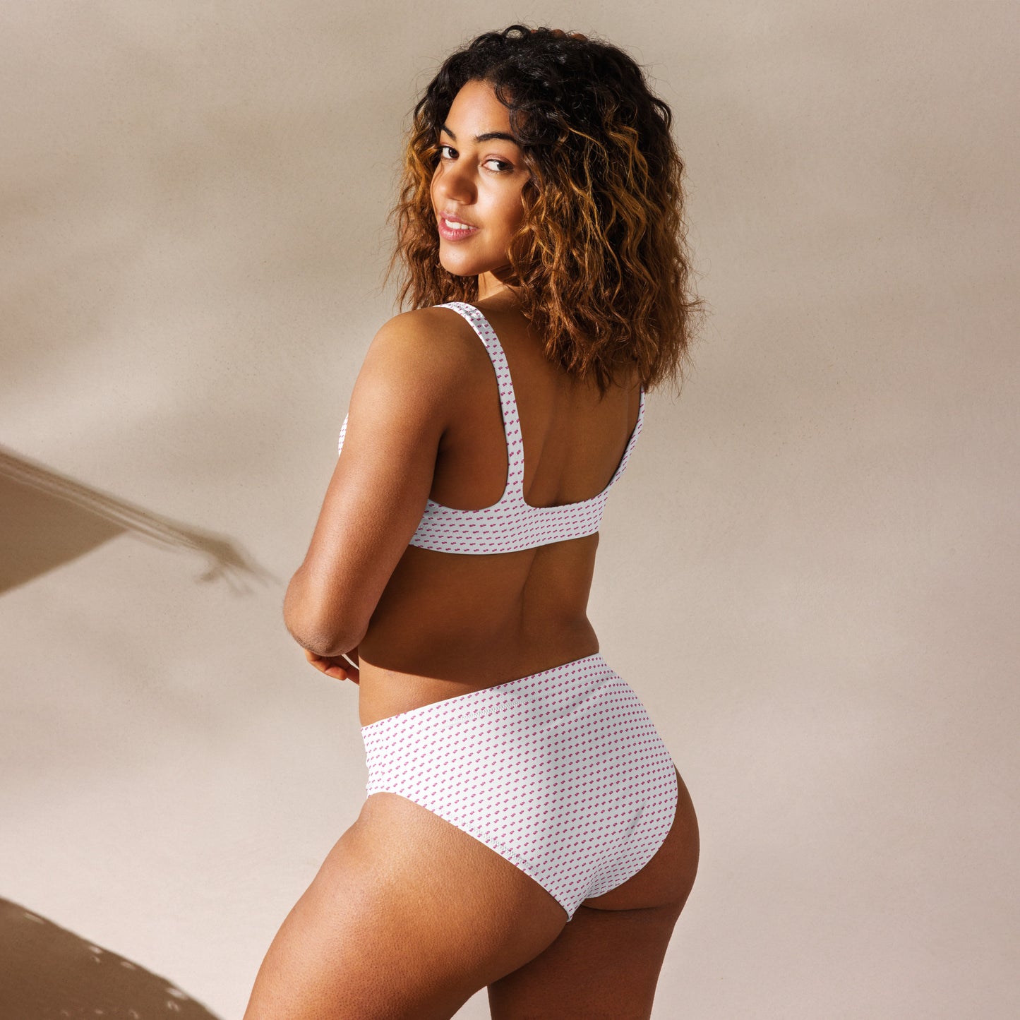 Curly-haired model in back-left view of slay bikini, white high-waisted bottoms and top with pink print