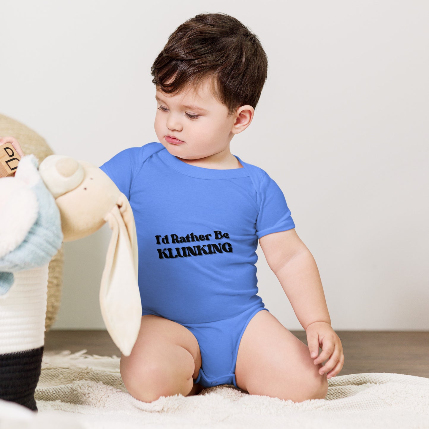 Baby cuddling teddy bear in light blue "I’d Rather Be Klunking" onesie, playful pout on