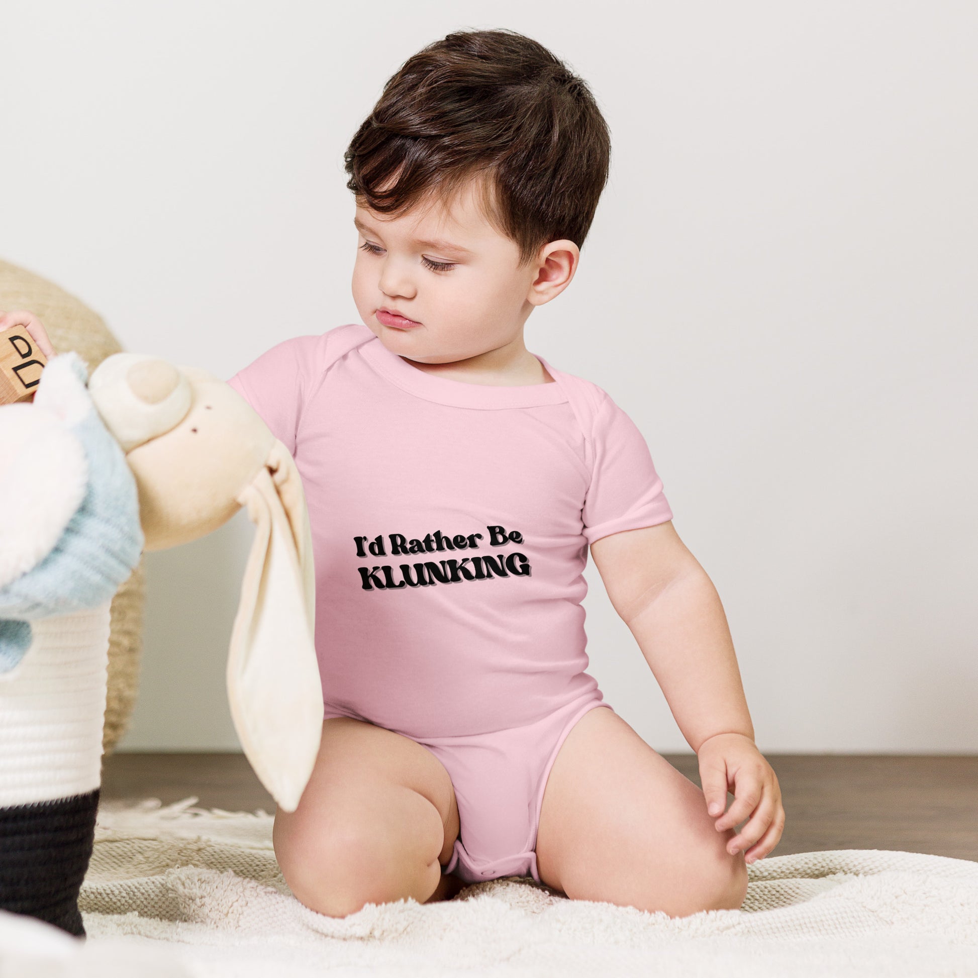 Thoughtful baby with teddy bear wearing pink "I’d Rather Be Klunking" onesie