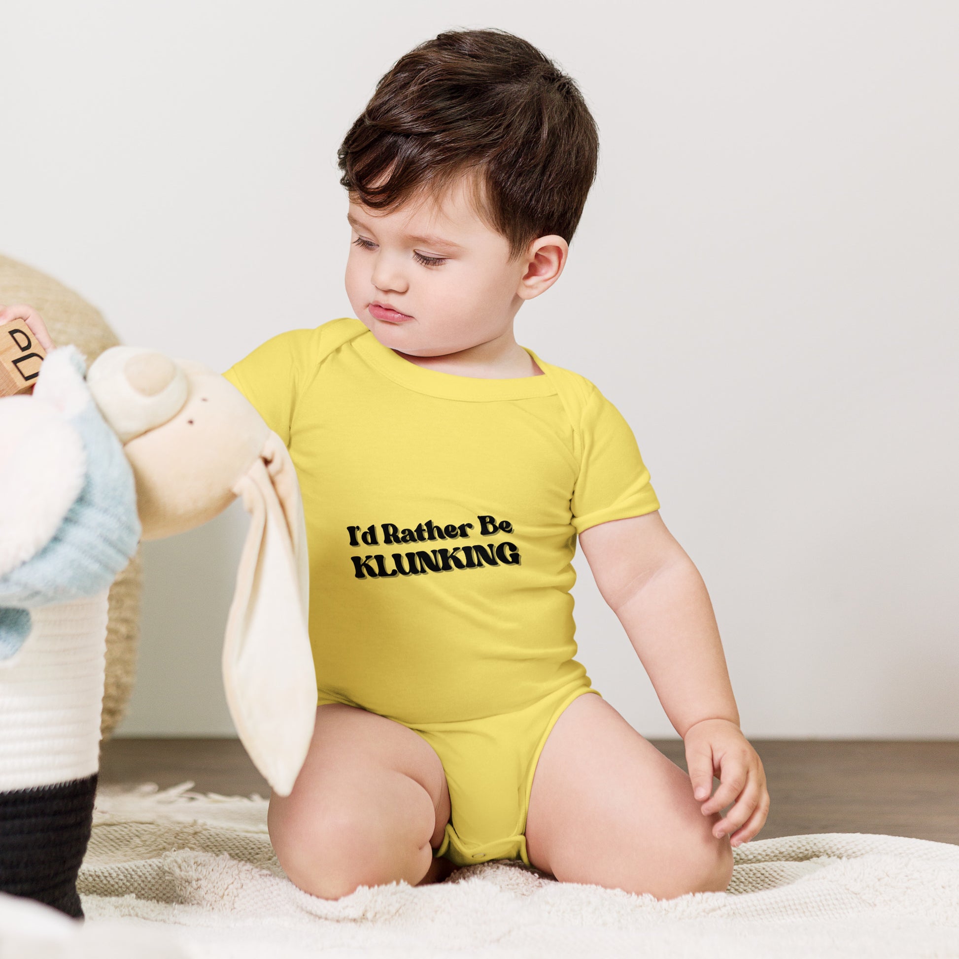 Baby clutching teddy in yellow "I’d Rather Be Klunking" onesie, adventure ready