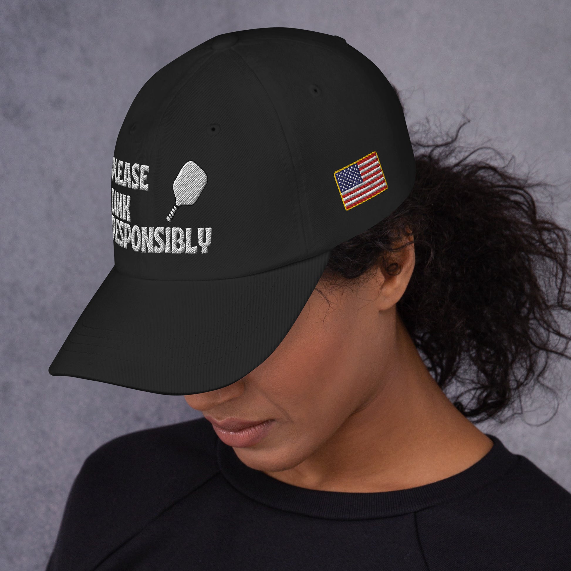 Left side profile of black dad hat on model, partial "PLEASE DINK RESPONSIBLY" embroidery, pickleball paddle, and small US flag patch