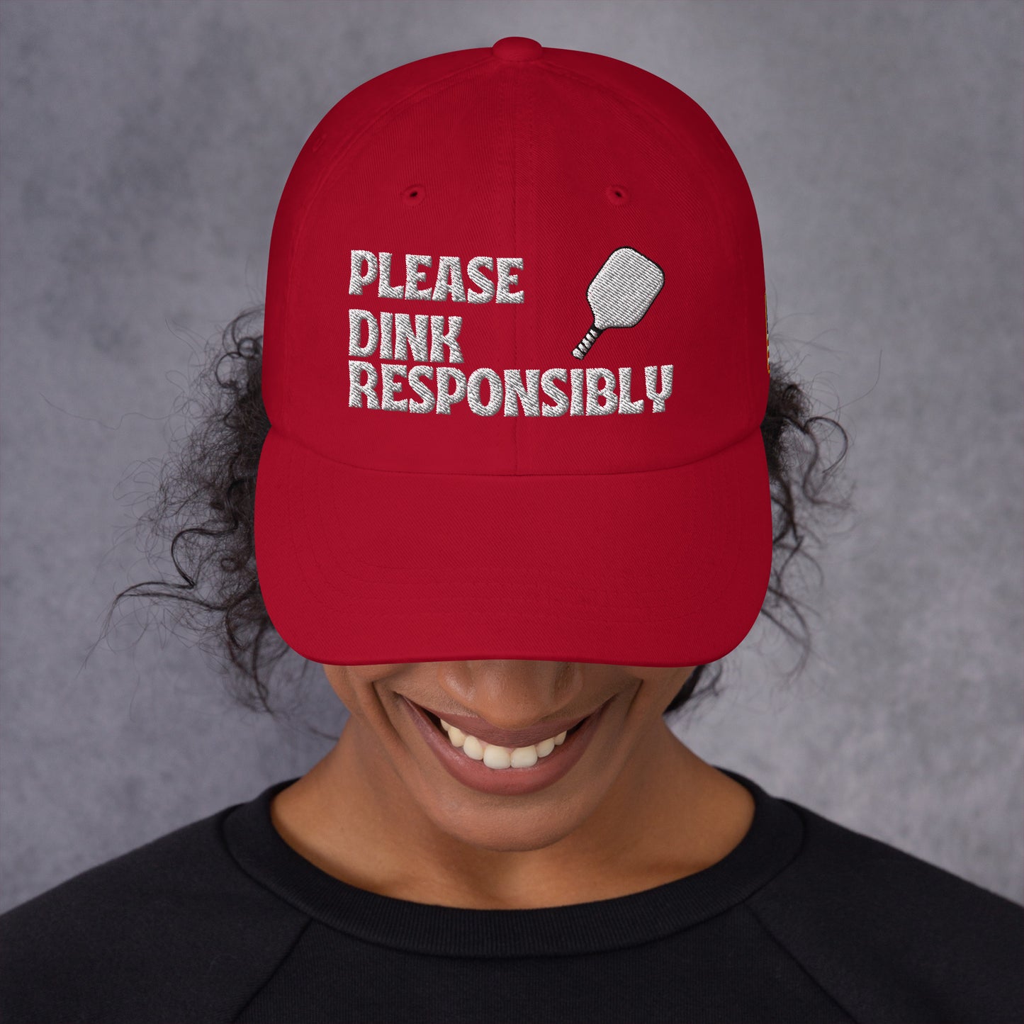 Front view of cranberry red dad hat on smiling model, white "PLEASE DINK RESPONSIBLY" embroidery with pickleball paddle icon