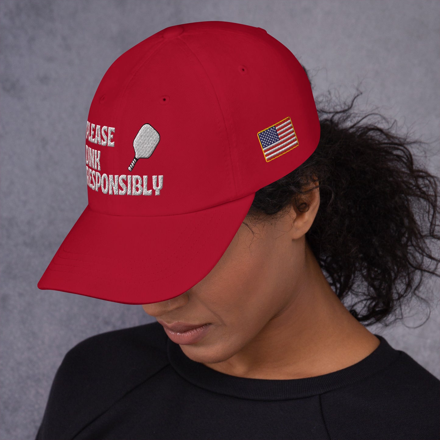 Left side profile of cranberry red dad hat on model, partial "PLEASE DINK RESPONSIBLY" embroidery, pickleball paddle, and small US flag patch