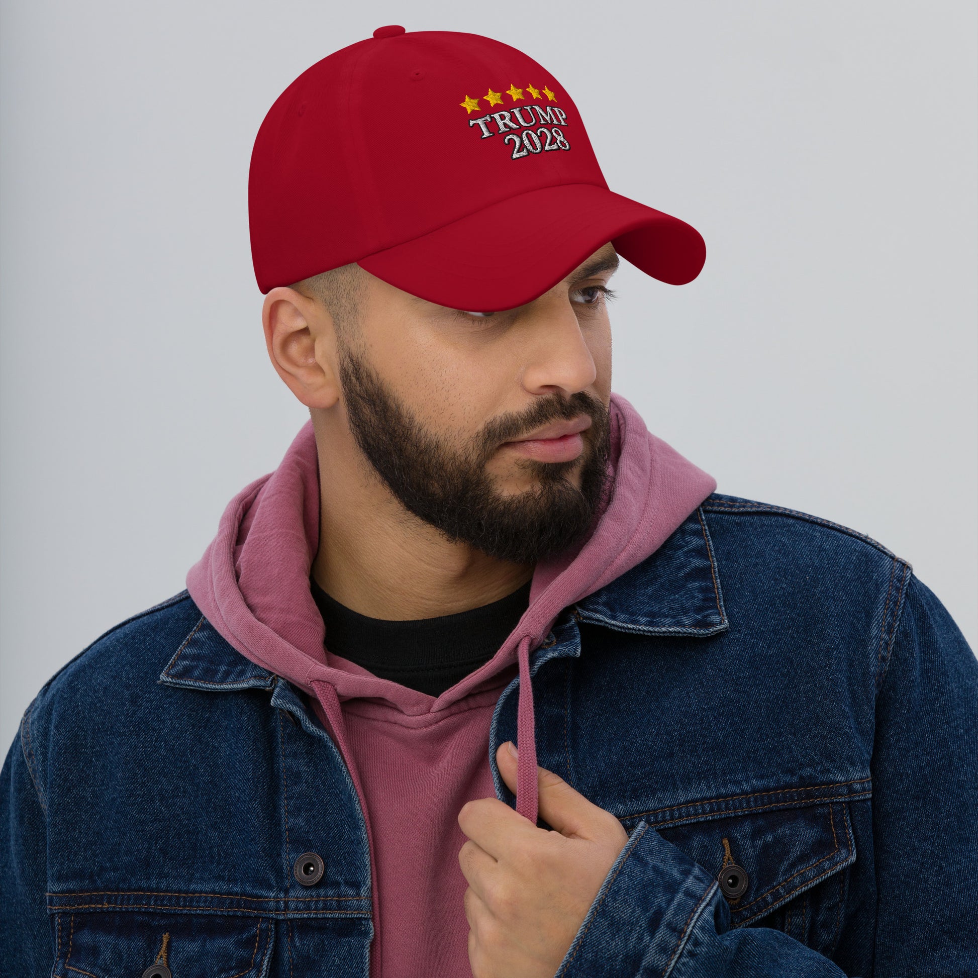 Profile of bearded man in hoodie wearing red dad hat, partial "TRUMP 2028" embroidery visible