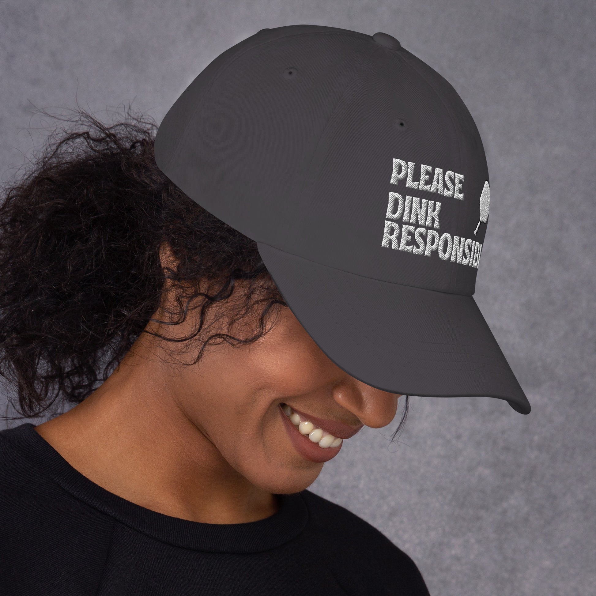 Model smiling in dark grey dad hat showing "PLEASE DINK RESPONSIBLY" embroidery and pickleball paddle