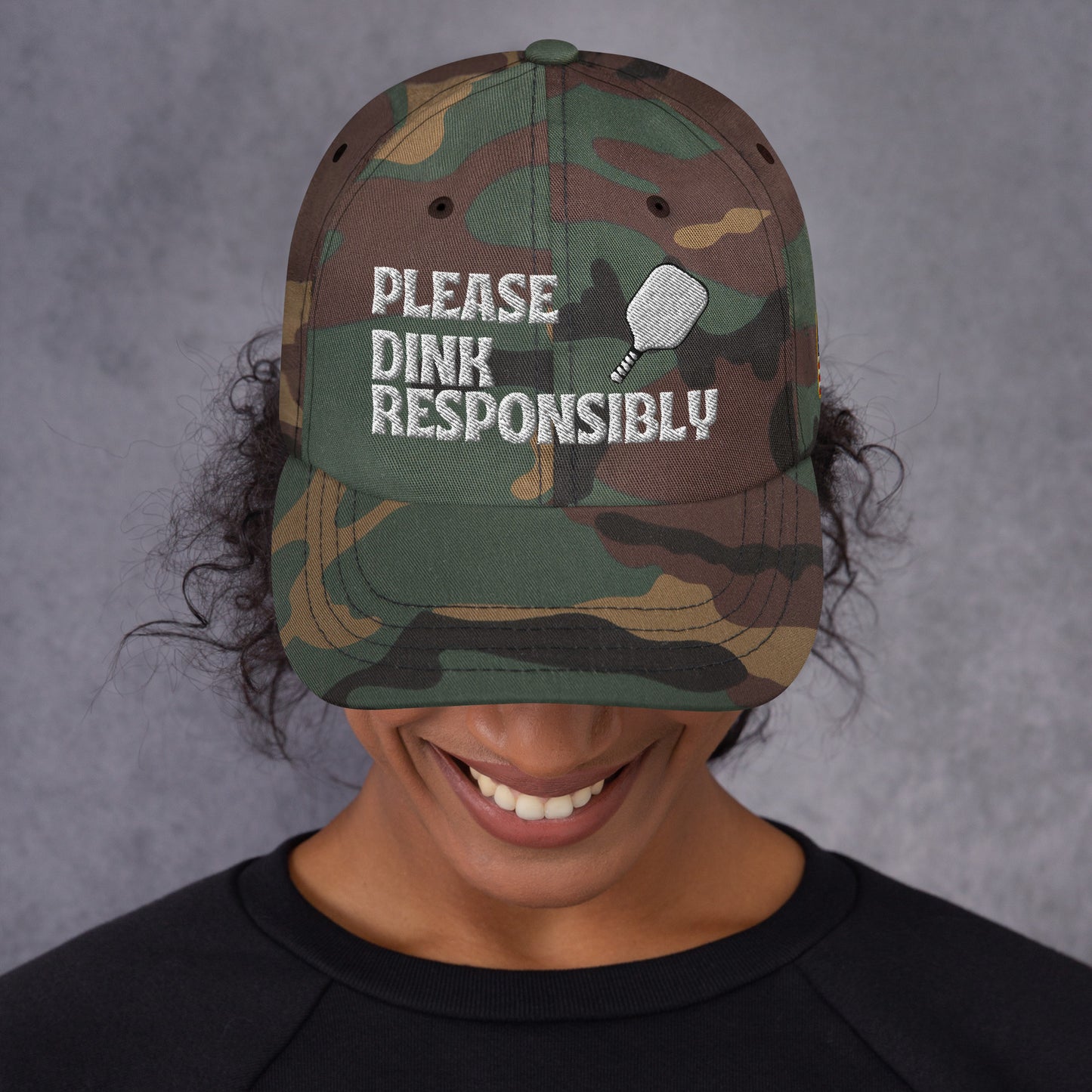 Front view under green camo dad hat on smiling model with "PLEASE DINK RESPONSIBLY" and pickleball embroidery