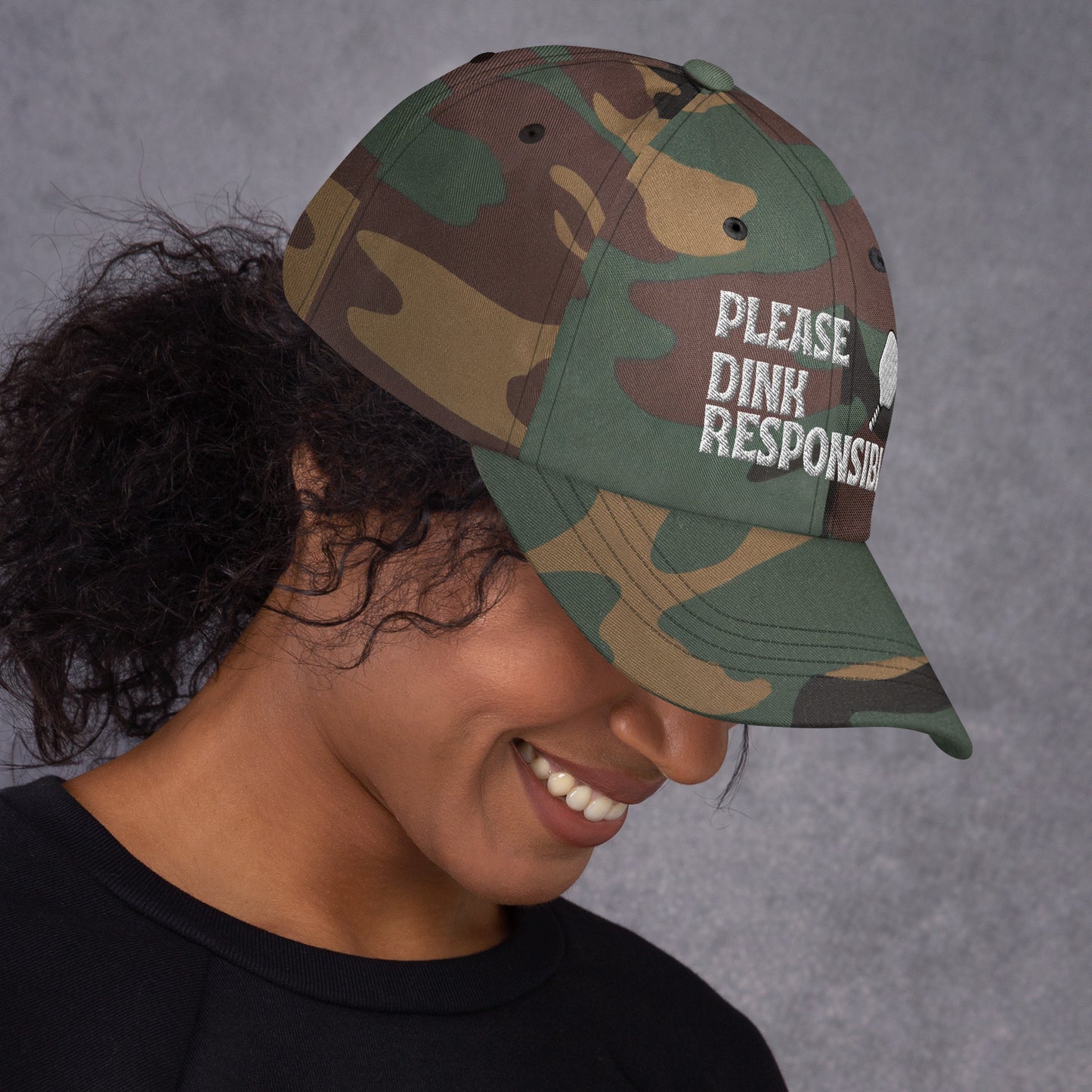 Smiling model under khaki dad hat revealing "PLEASE DINK RESPONSIBLY" embroidery and pickleball paddle
