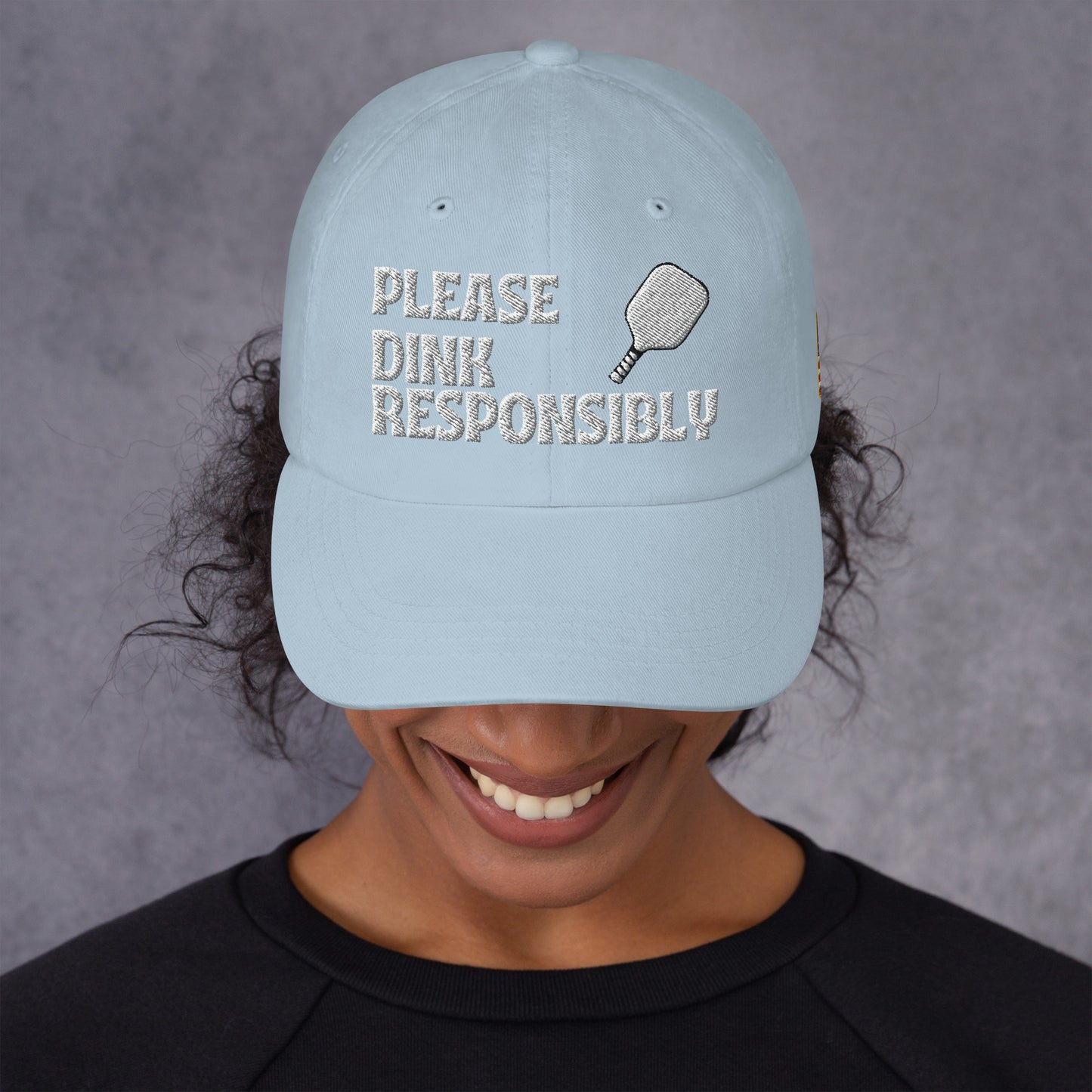 Light blue dad hat on grinning model, full "PLEASE DINK RESPONSIBLY" text with pickleball paddle embroidery