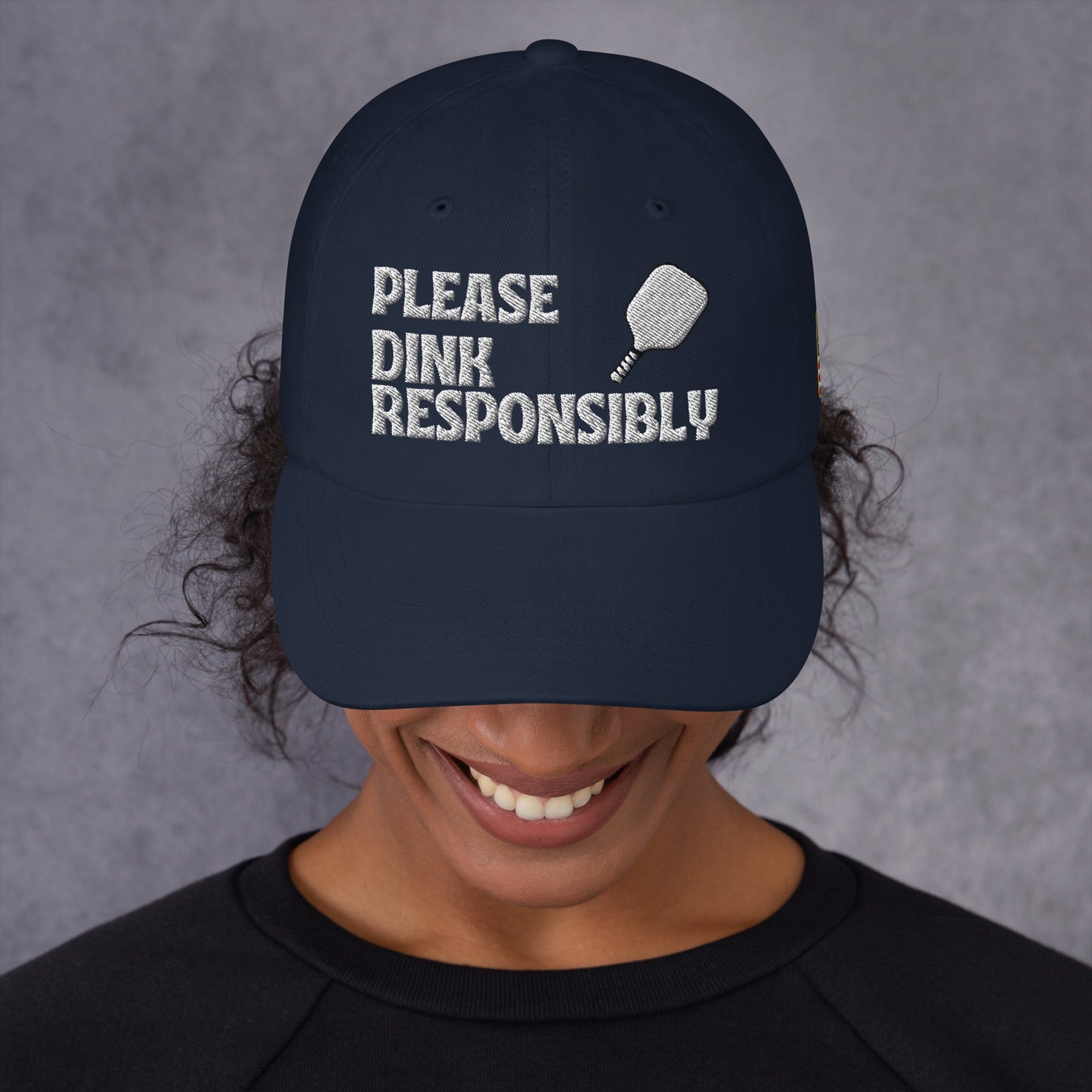 Front view of navy dad hat on smiling model, white "PLEASE DINK RESPONSIBLY" embroidery with pickleball paddle icon