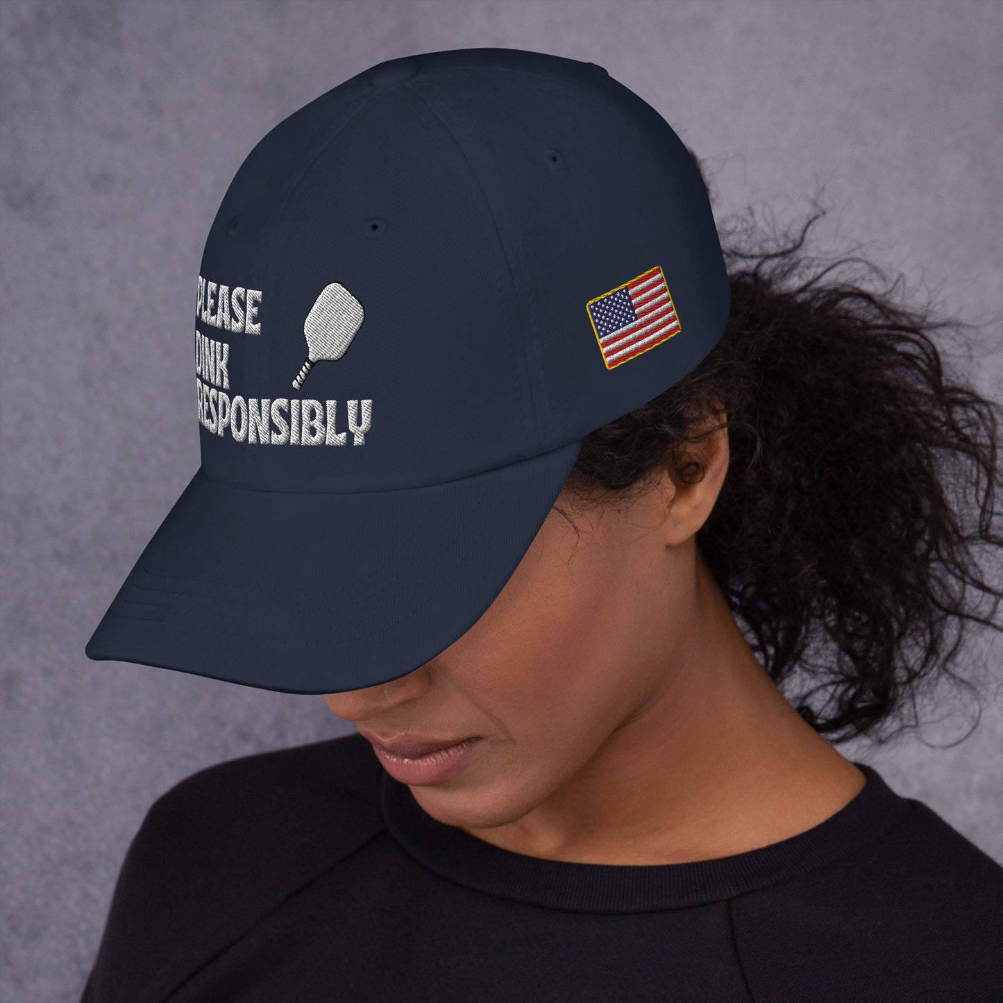 Left side profile of navy dad hat on model, partial "PLEASE DINK RESPONSIBLY" embroidery, pickleball paddle, and small US flag patch