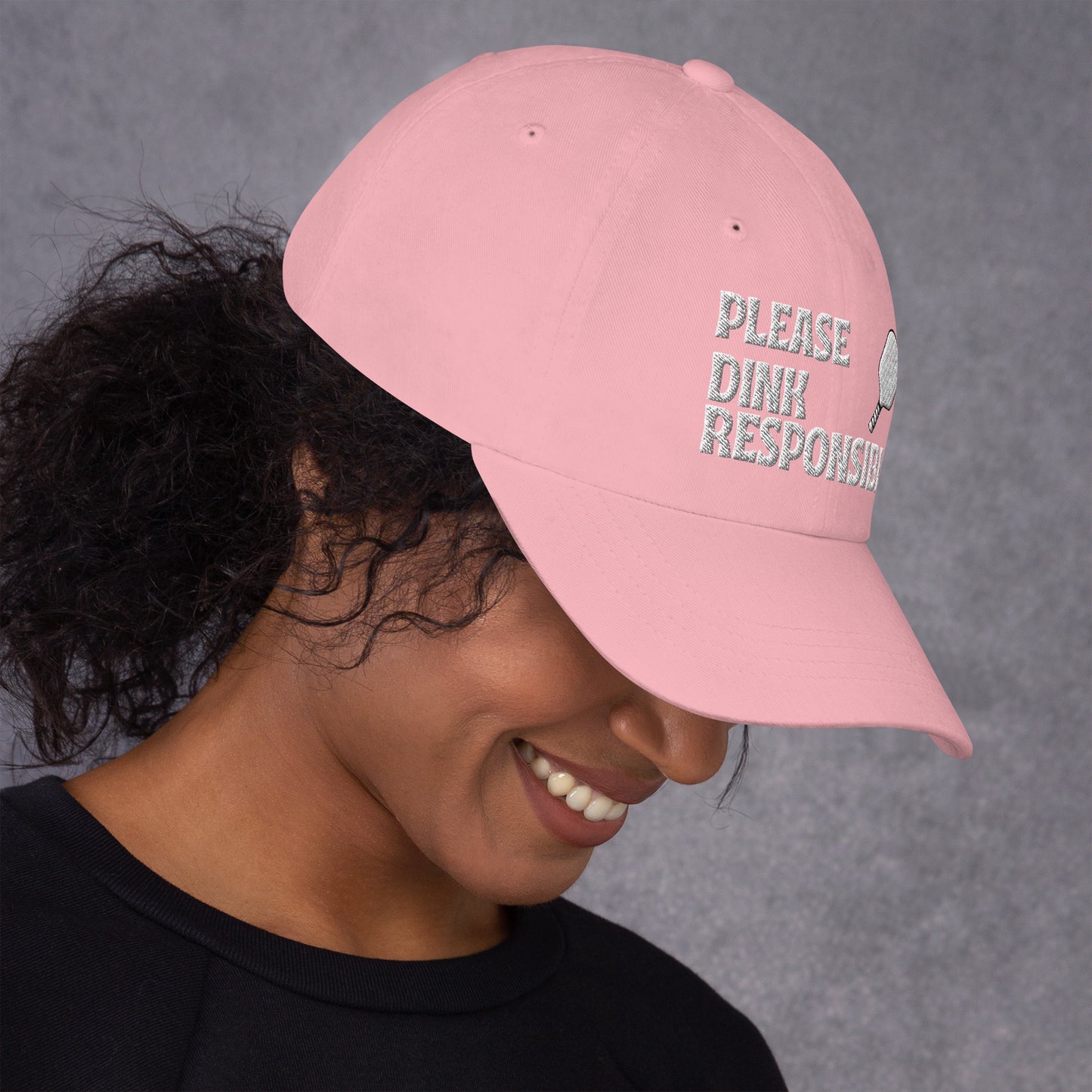 Smiling model in pink dad hat showing "PLEASE DINK RESPONSIBLY" arched embroidery and pickleball paddle