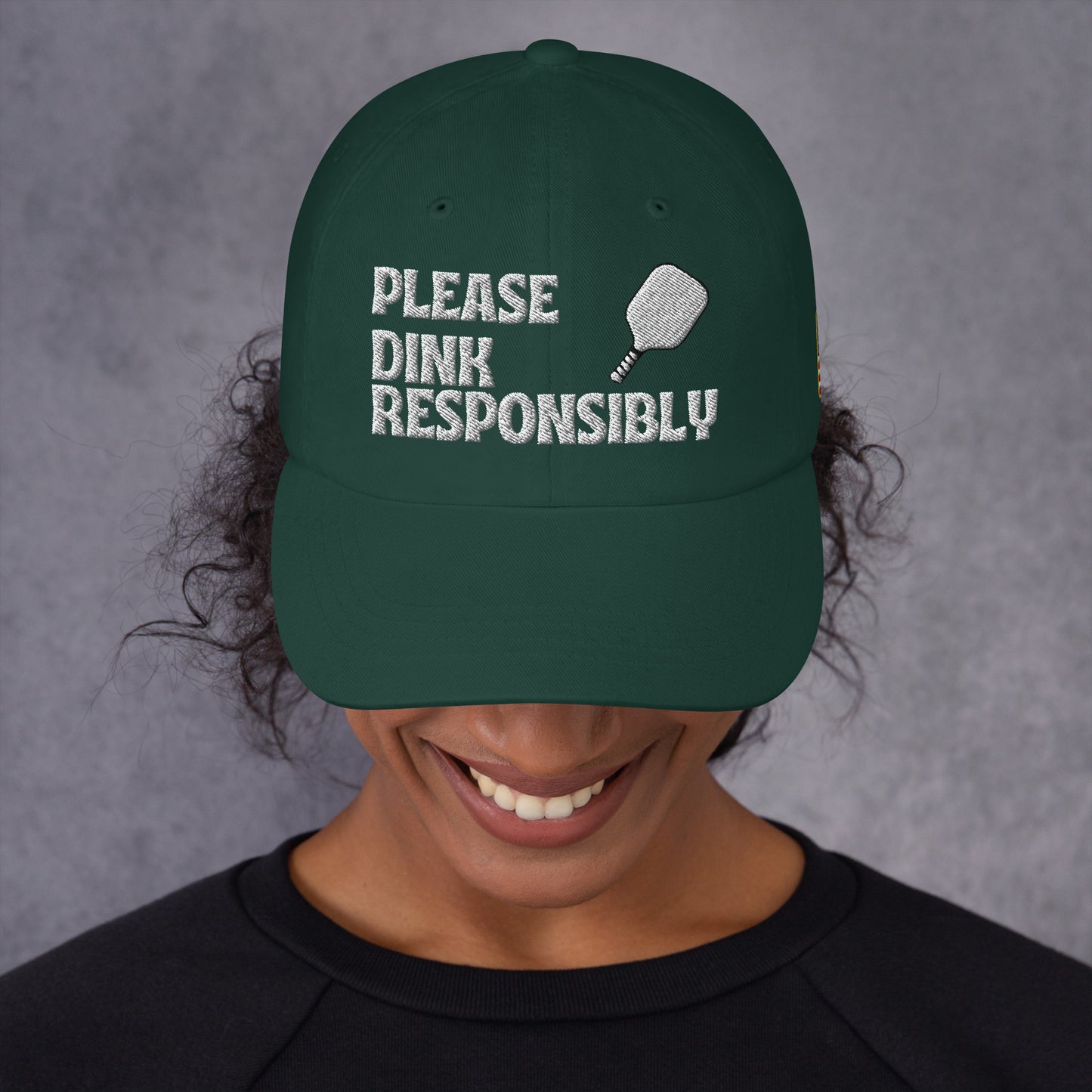 Front view of spruce green dad hat on smiling model, white "PLEASE DINK RESPONSIBLY" embroidery with pickleball paddle icon