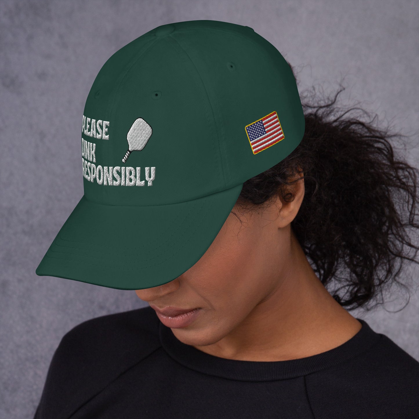 Left side profile of spruce green dad hat on model, partial "PLEASE DINK RESPONSIBLY" embroidery, pickleball paddle, and small US flag patch