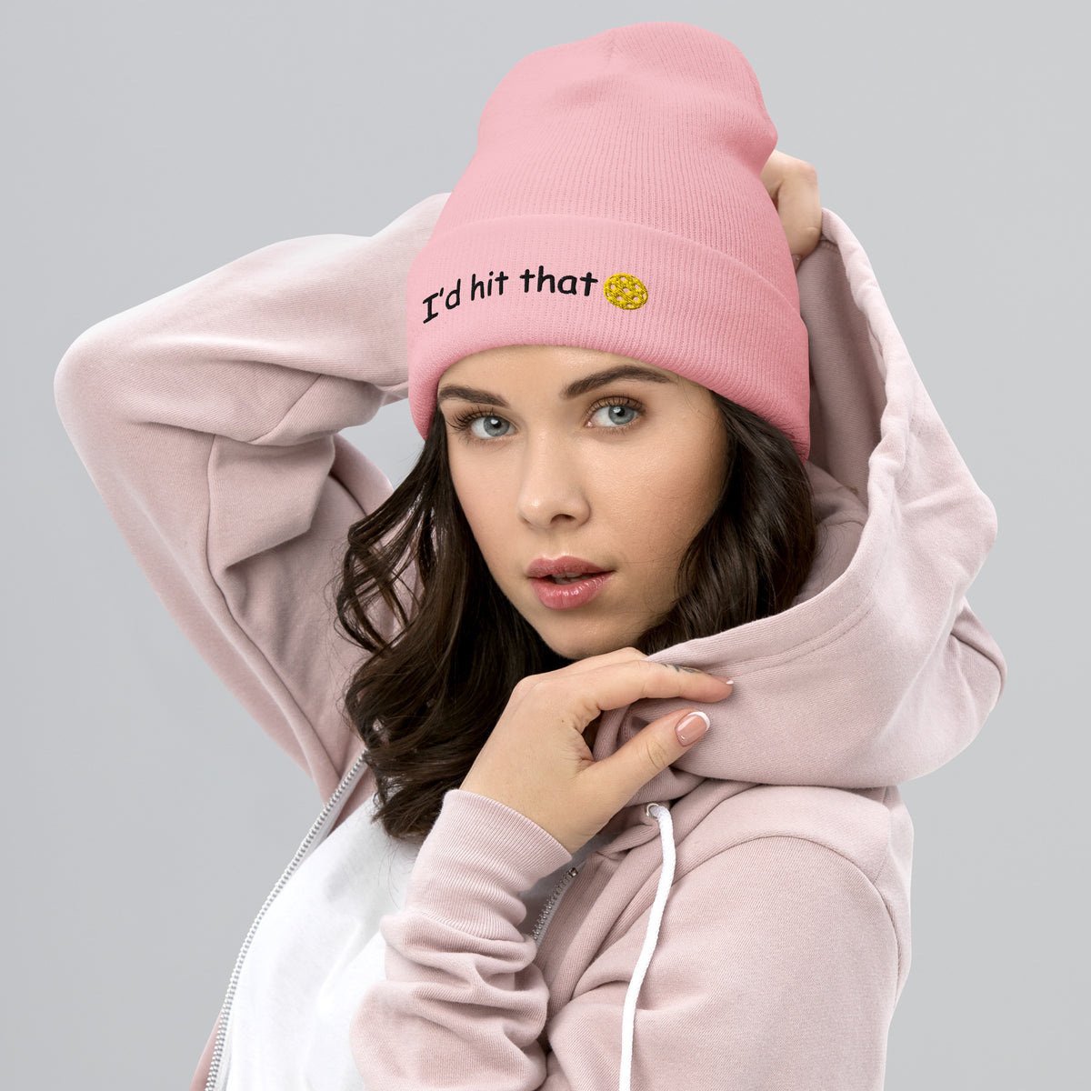 Model in playful pose wearing baby pink cuffed beanie with yellow "I'd hit that 🥒" embroidery, arms raised over grey hoodie