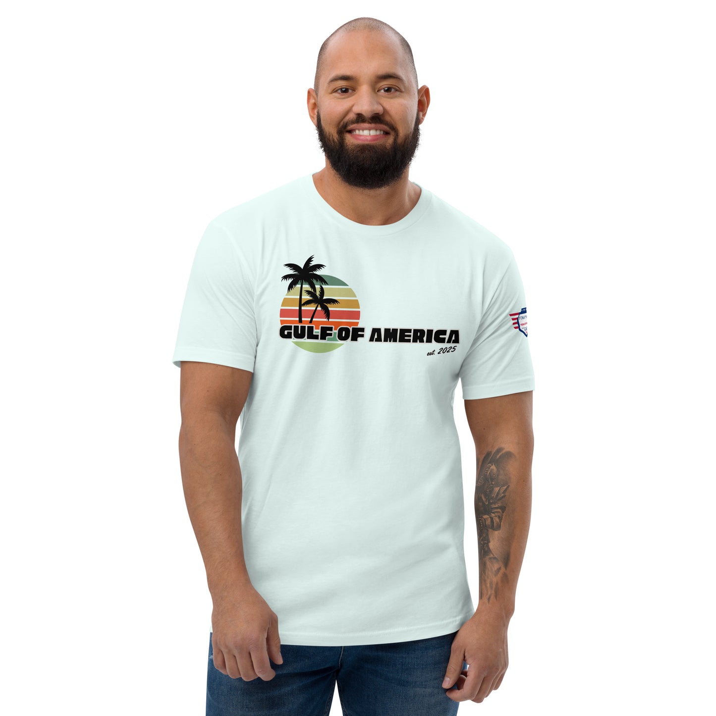 Smiling bearded model in front view of light blue fitted tee with retro black/orange palm sunset graphic, arched "GULF OF AMERICA" text, and USA flag sleeve patch