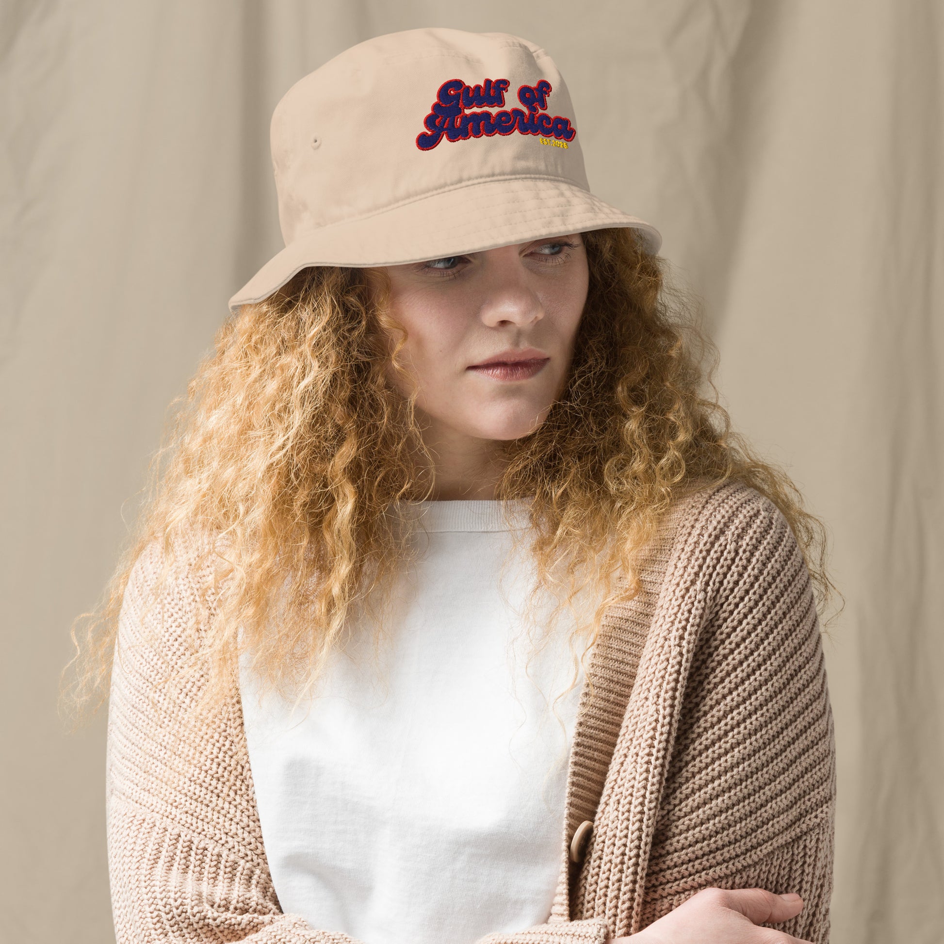Curly-haired model in three-quarter view wearing beige bucket hat, "Gulf of America" multicolored print front and center