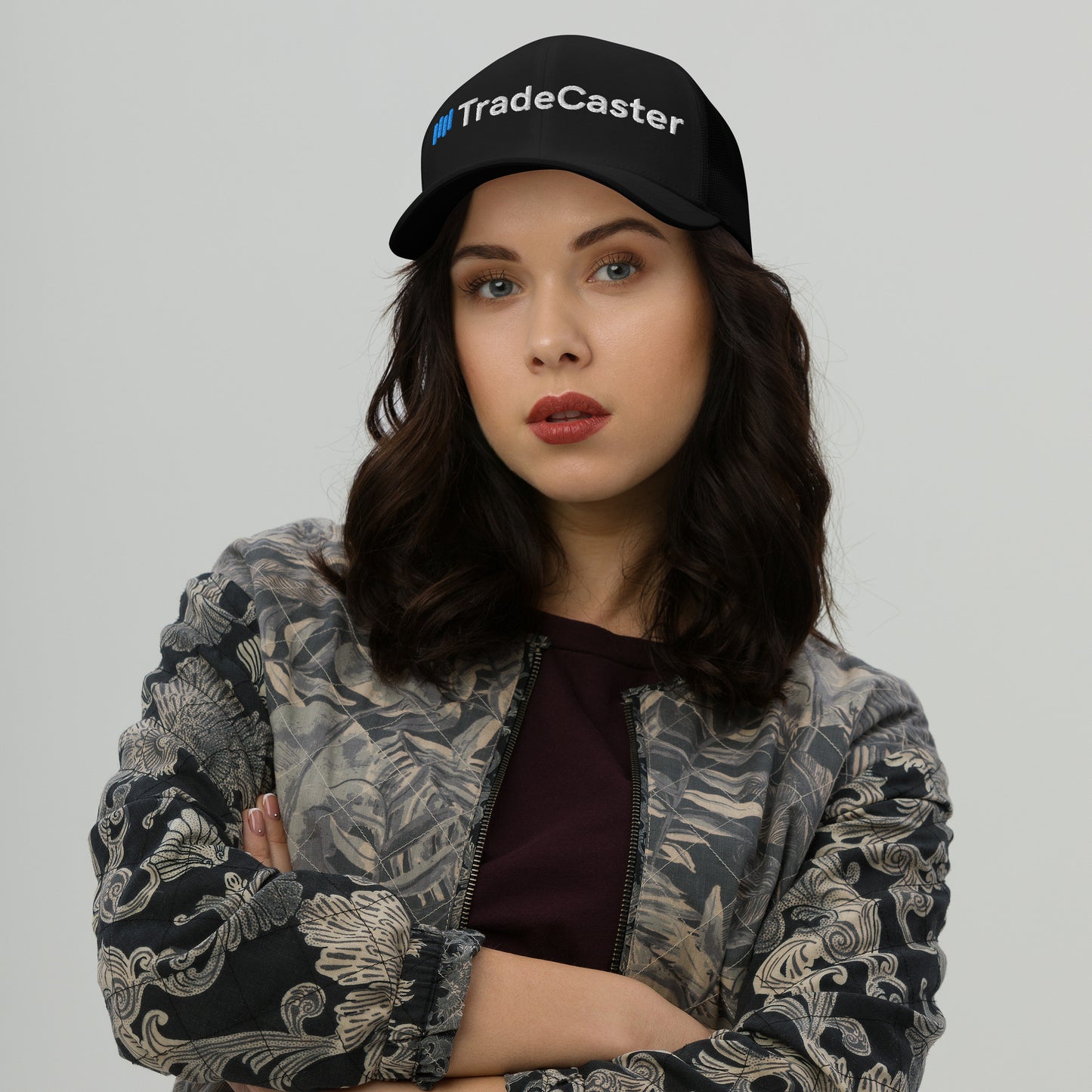 Woman model rocking black TradeCaster trucker cap front view, blue/white embroidery pops, arms crossed vibe