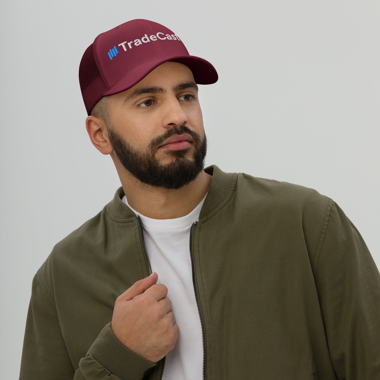 Bearded guy in burgundy TradeCaster trucker cap, side profile view, white mesh back, green bomber jacket — ready to sling trades