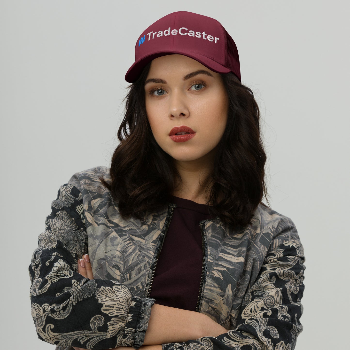 Woman with wavy hair rocking burgundy TradeCaster trucker cap, attitude-forward pose, arms crossed in patterned jacket