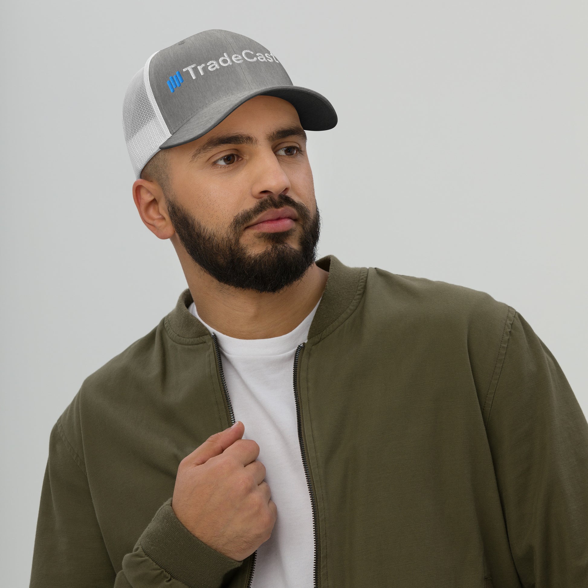 Bearded model sporting gray TradeCaster trucker cap from side, white mesh back breezy, green jacket vibes