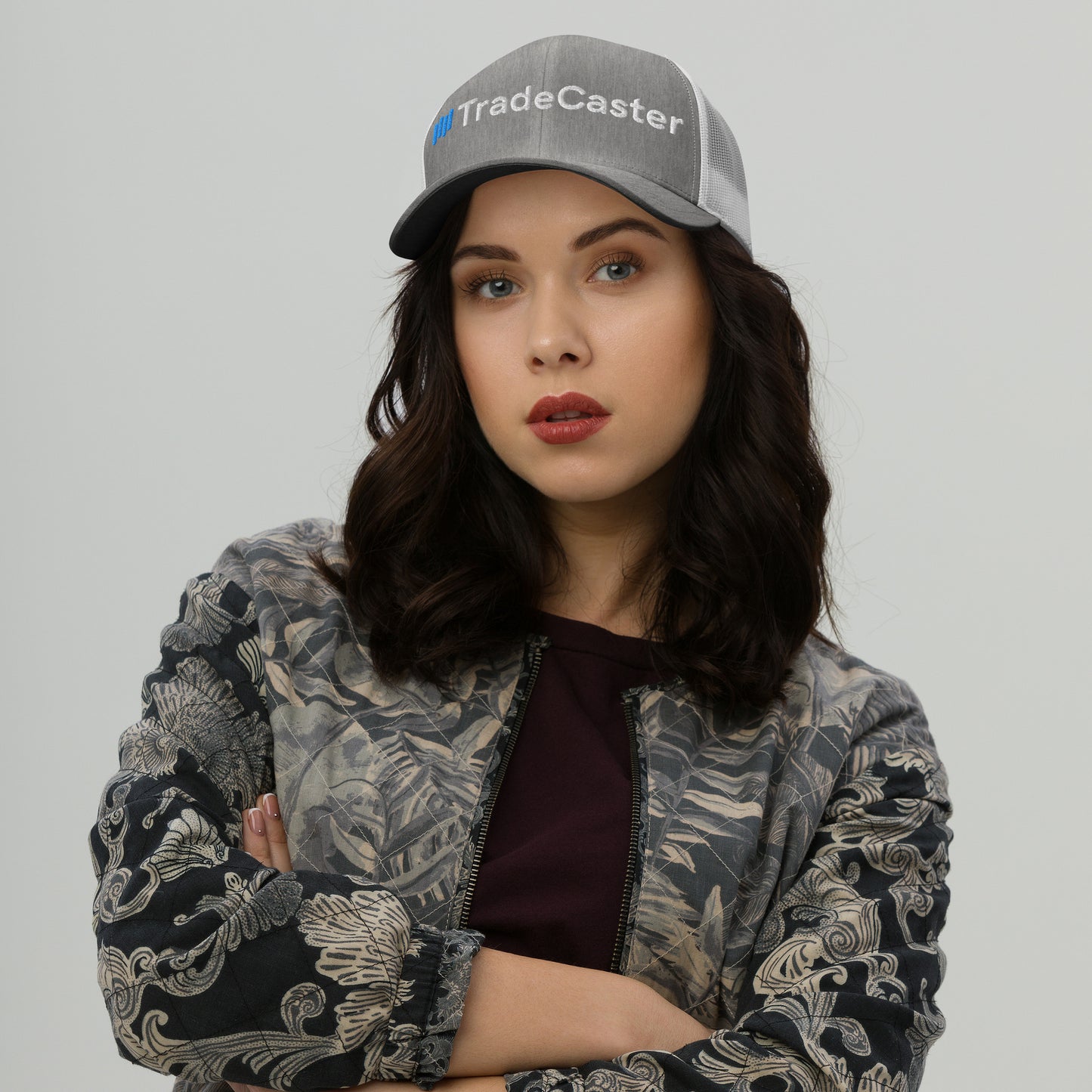 Dark-haired model in gray TradeCaster trucker cap, sassy front stance, arms crossed over patterned jacket